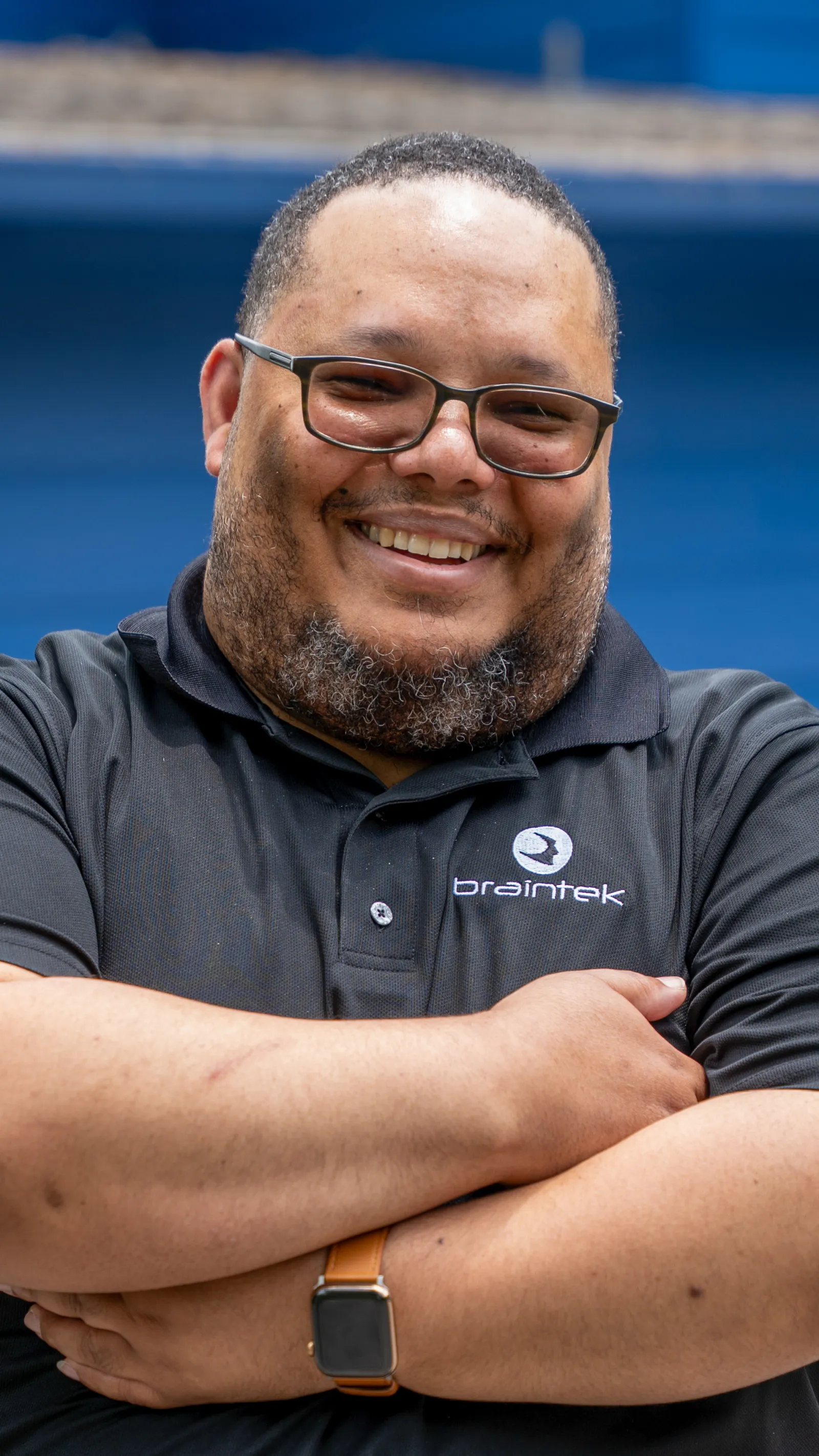 Smiling man wearing glasses and a Braintek polo shirt with arms crossed against a blue background.