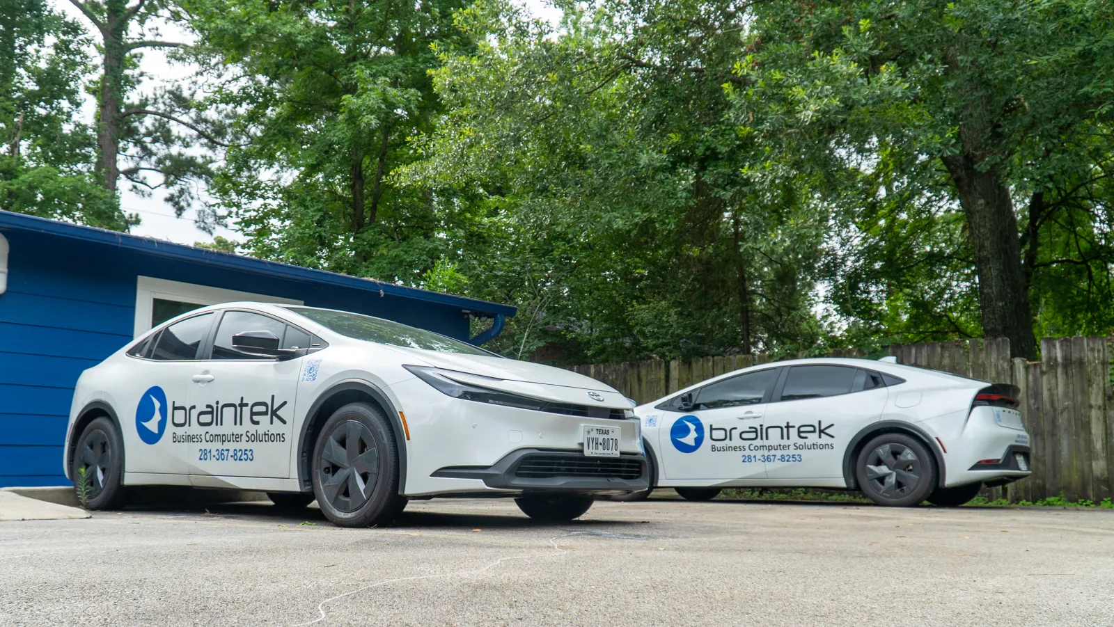 Two white Braintek business cars parked outside by blue building and green trees in daylight
