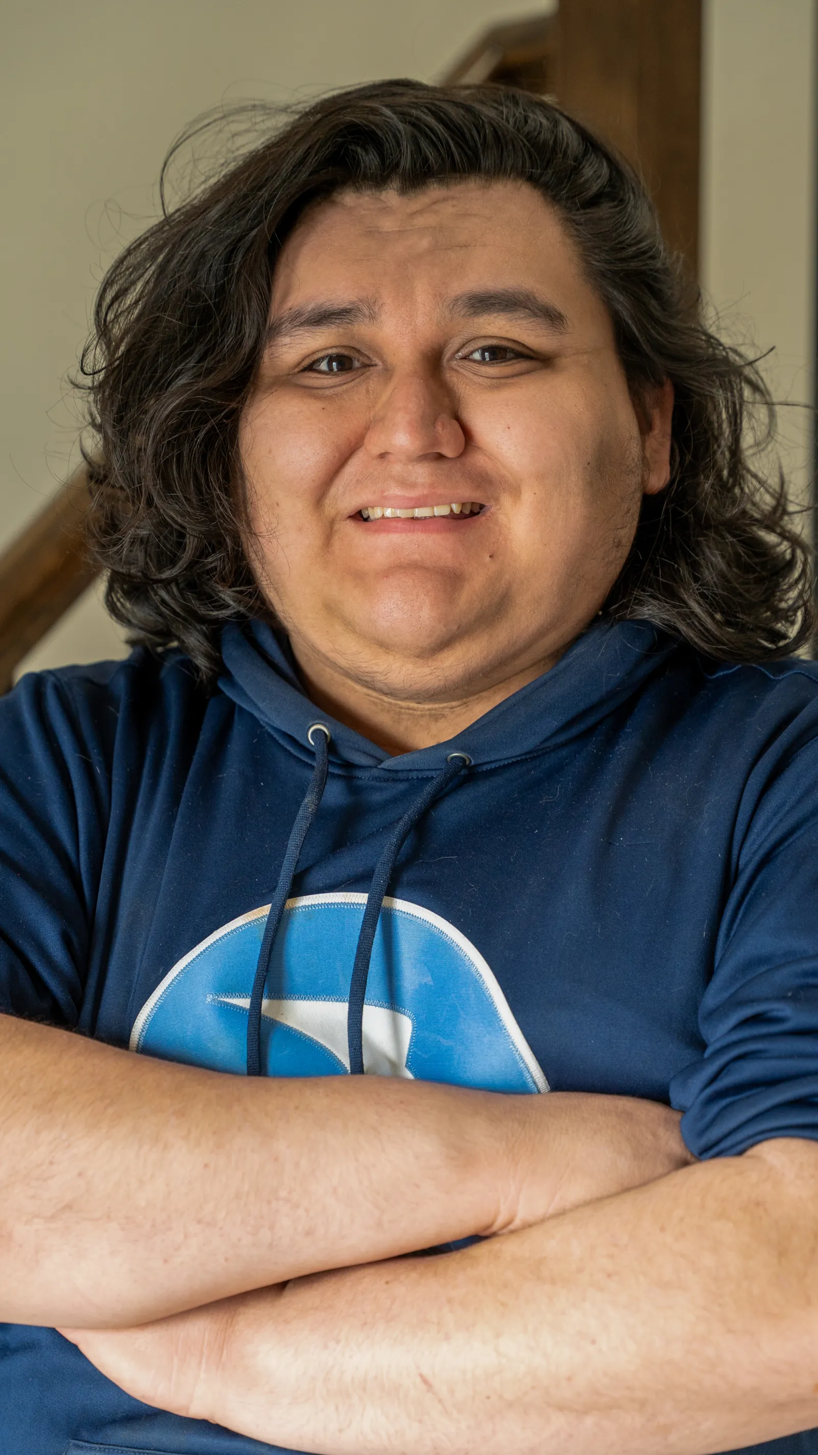 Smiling young man with long dark hair wearing a blue hoodie stands with arms crossed indoors near stairs.