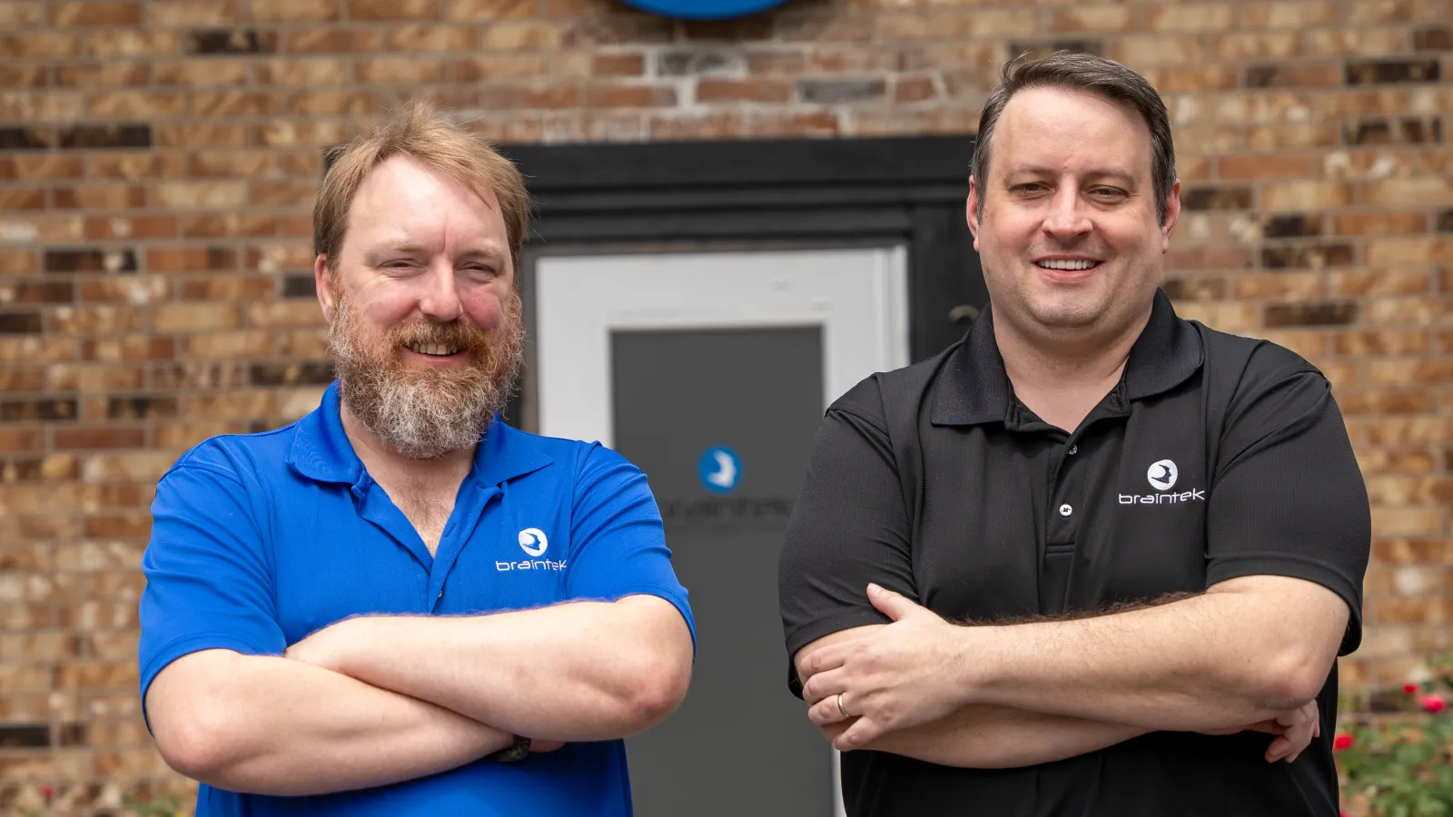 Two smiling men in branded polo shirts standing with arms crossed outside a brick building with company logo.