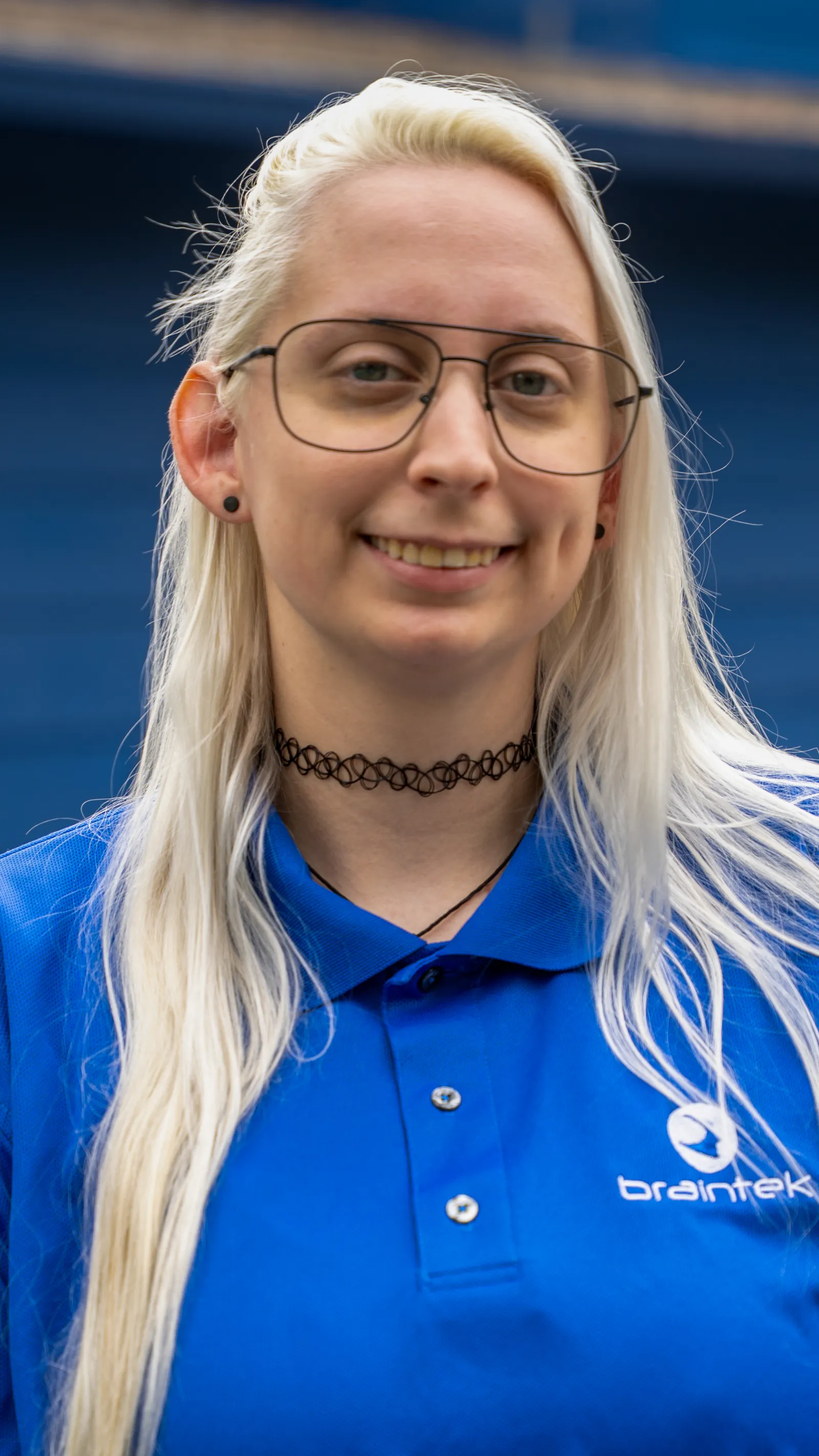 Young woman with long blonde hair in glasses wearing a blue Braintek polo shirt smiling outdoors.