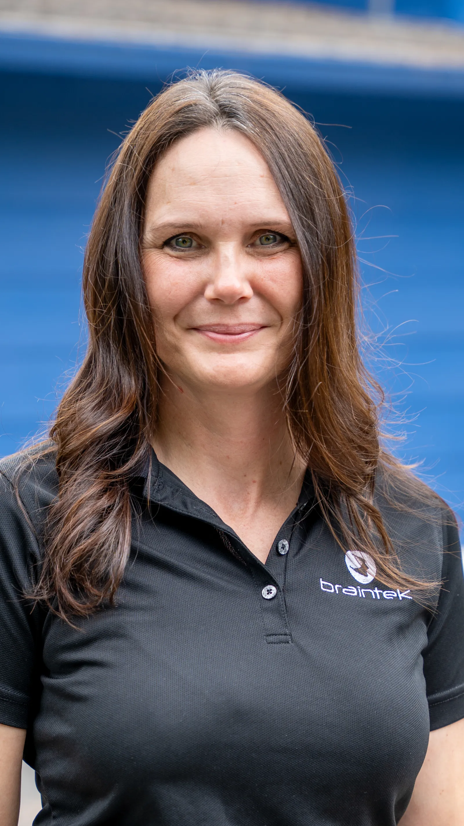 Smiling woman with long brown hair wearing a black Braintek polo shirt with a blue background.