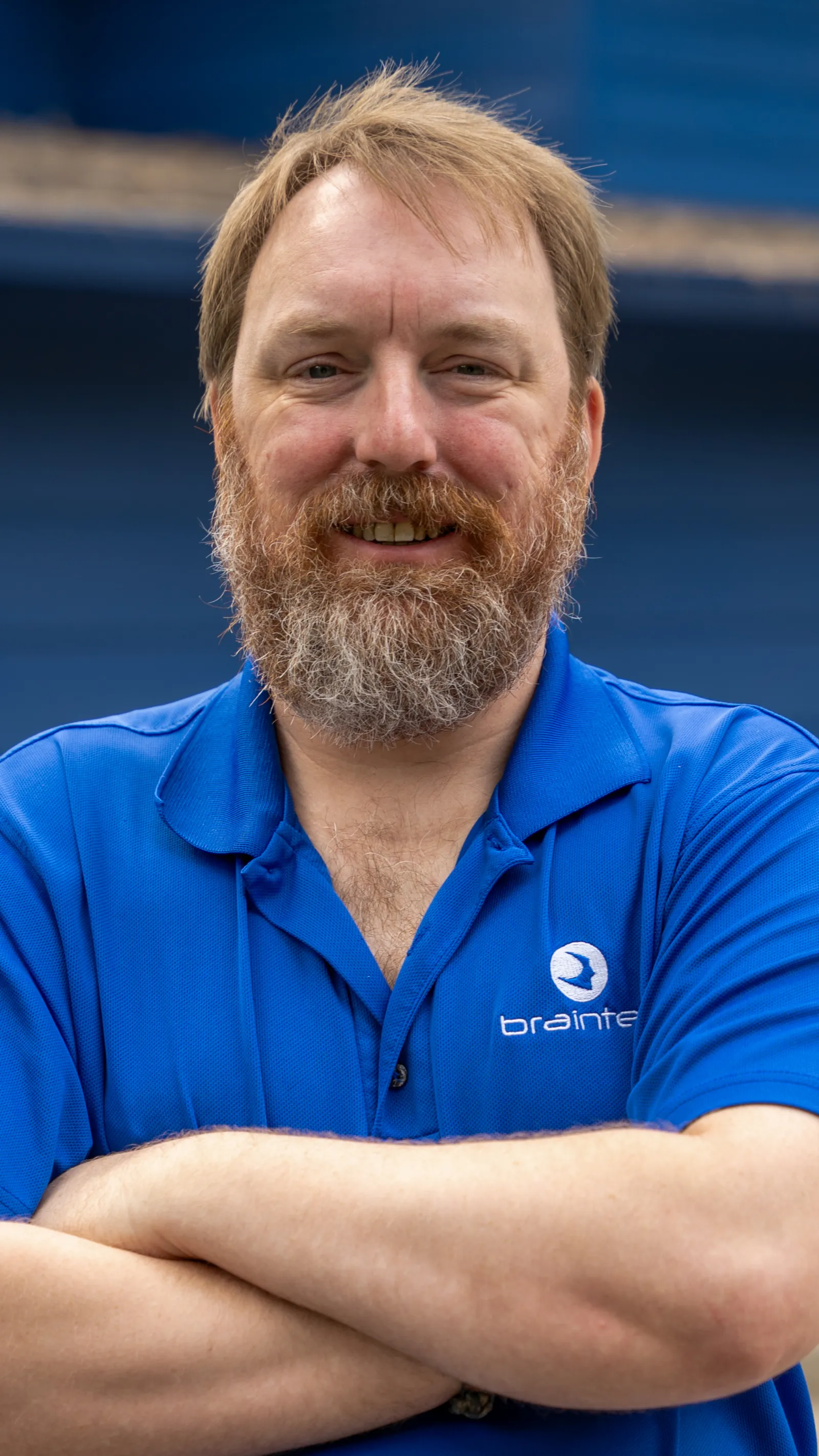 Smiling man with a beard wearing a blue Braintree polo shirt stands with arms crossed against a blue background.