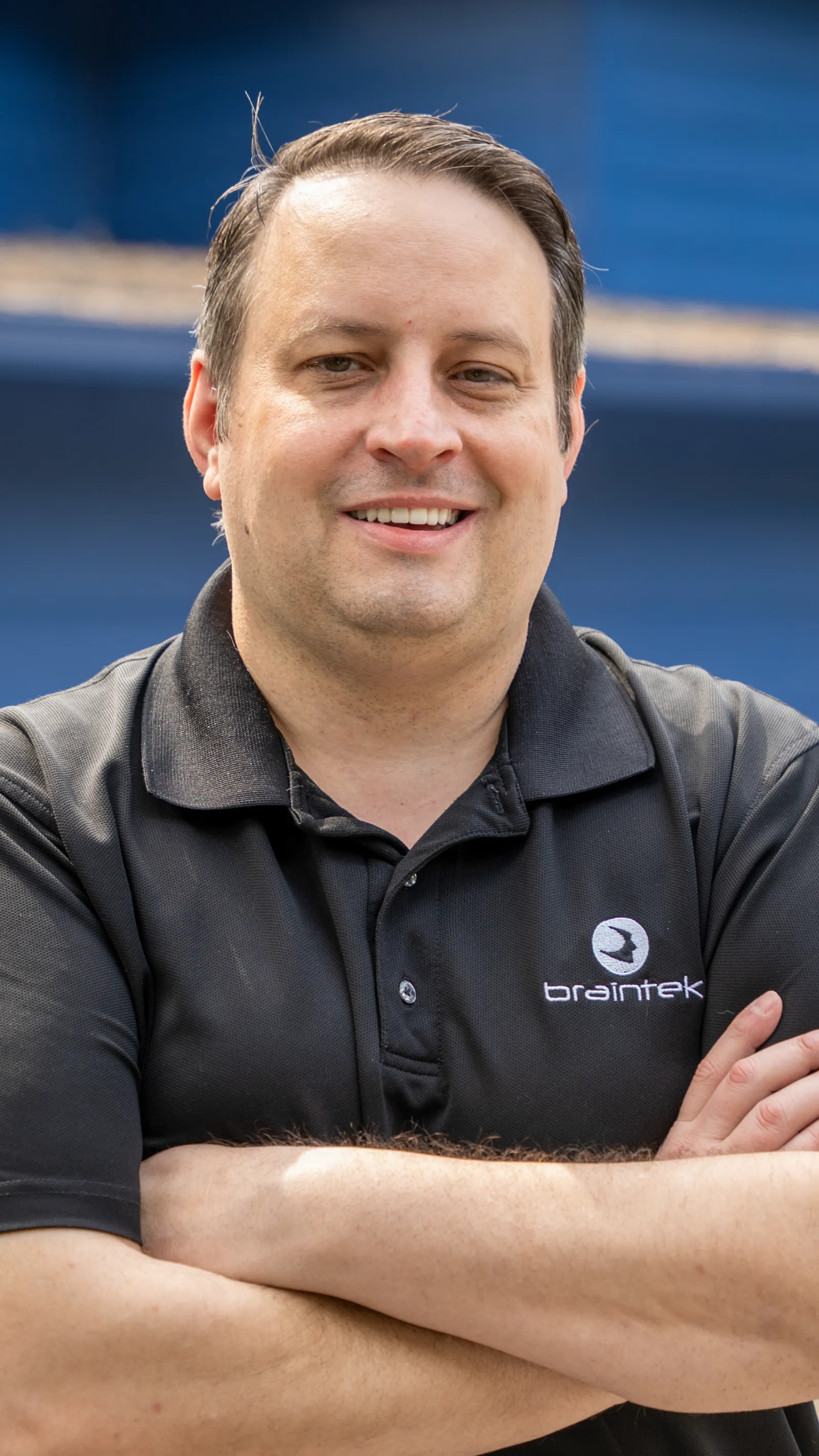 Smiling man in black Braintek polo shirt with arms crossed against blue background.