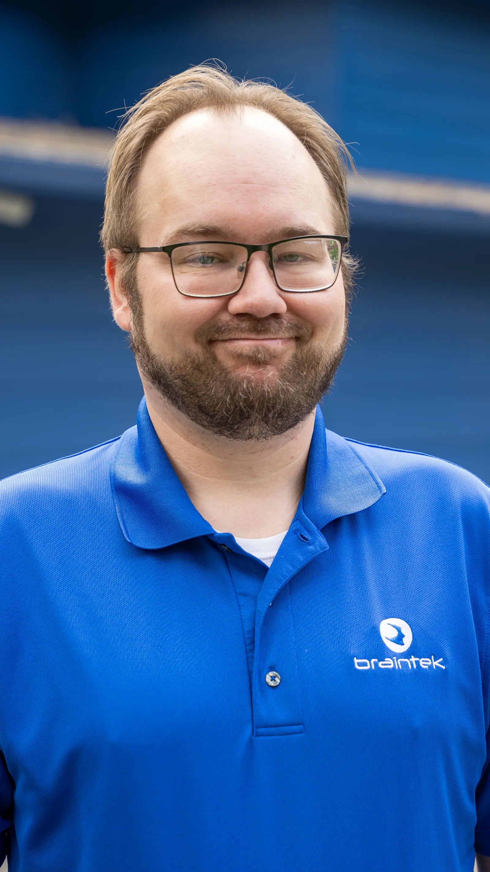 Smiling man with glasses and beard wearing a blue Braintek polo shirt outdoors with a blurred blue background.