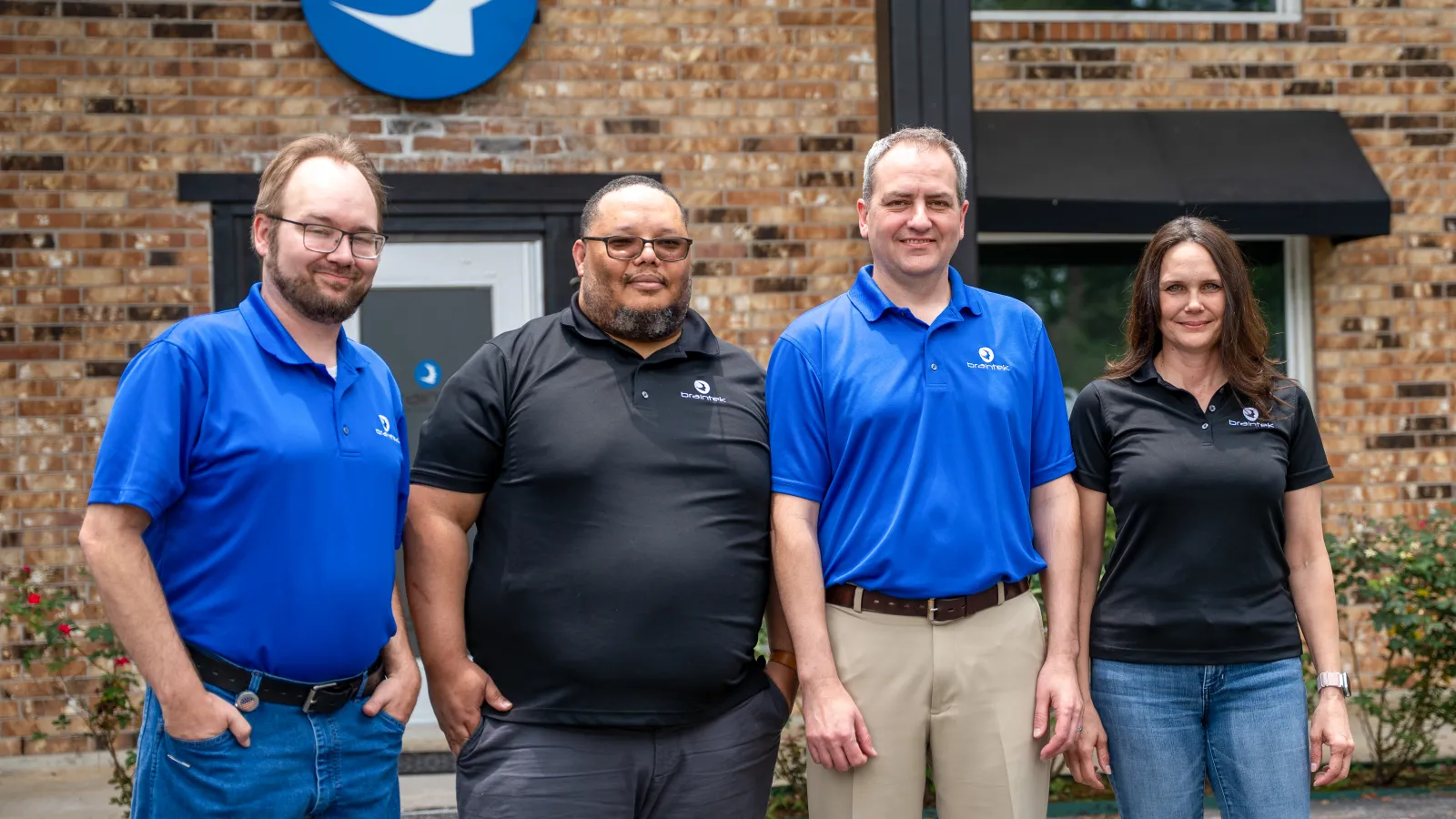 Four employees in blue and black company polo shirts standing outdoors in front of a brick building with a blue logo on the wall.