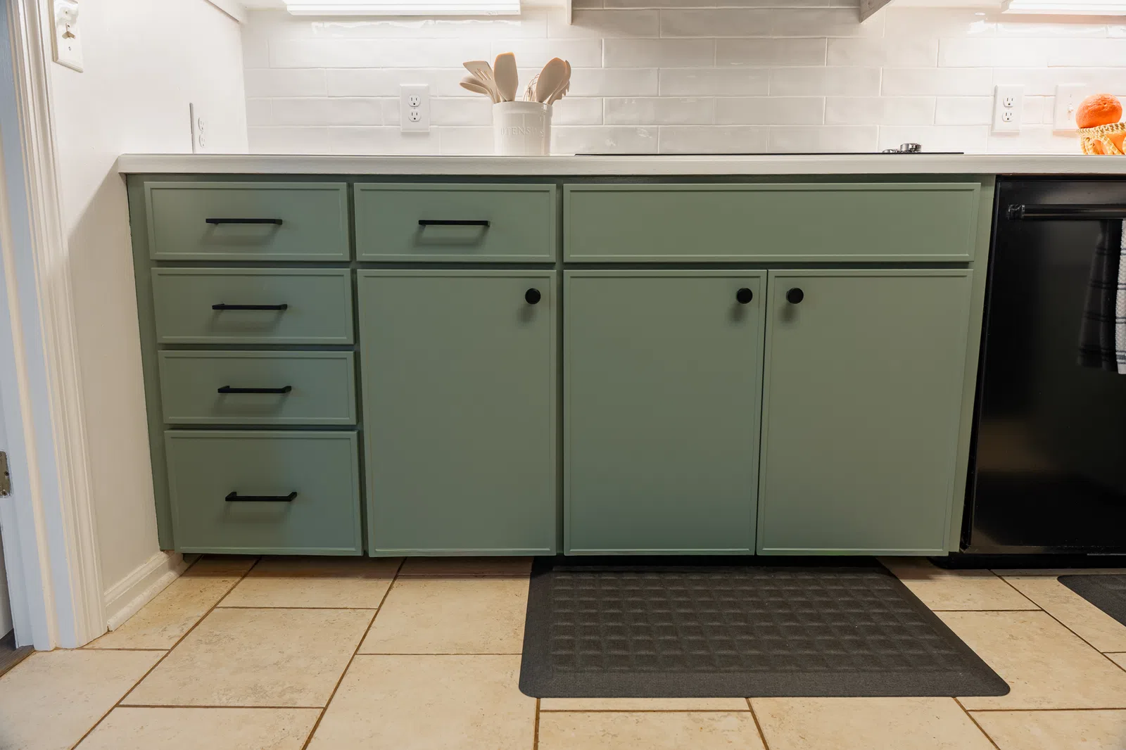 Light green kitchen cabinets with black handles and knobs below white countertop and tiled backsplash