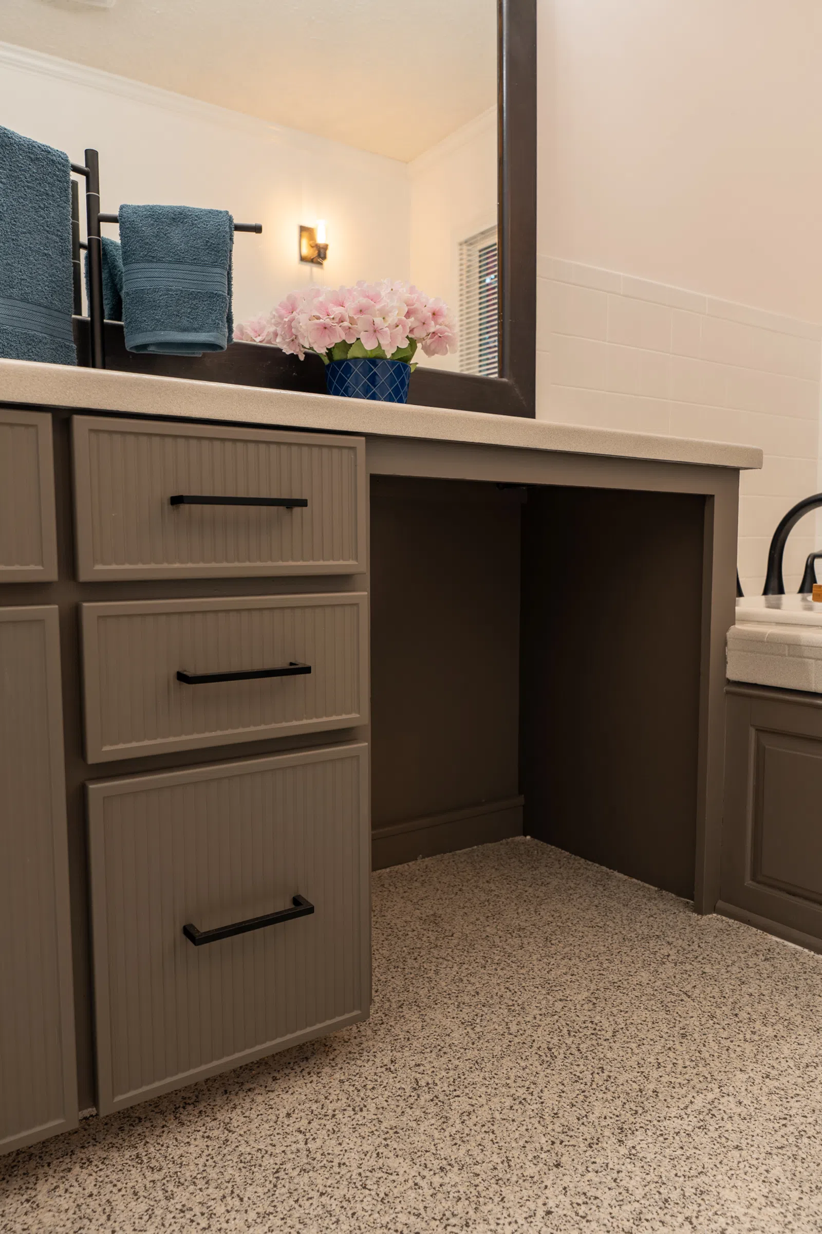 Brown bathroom vanity with black handles, pink flowers in blue vase, blue towels, and terrazzo floor.