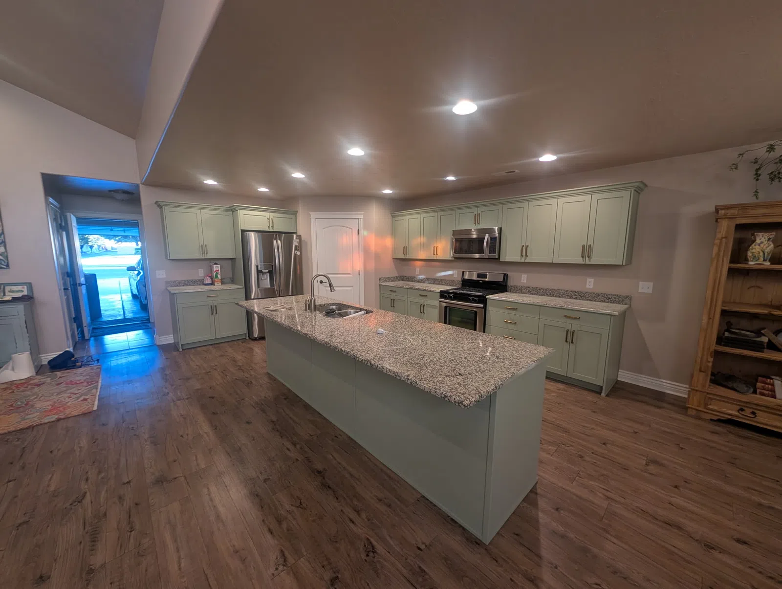 Modern kitchen with large granite island, light green cabinets, stainless steel appliances, and wood flooring.