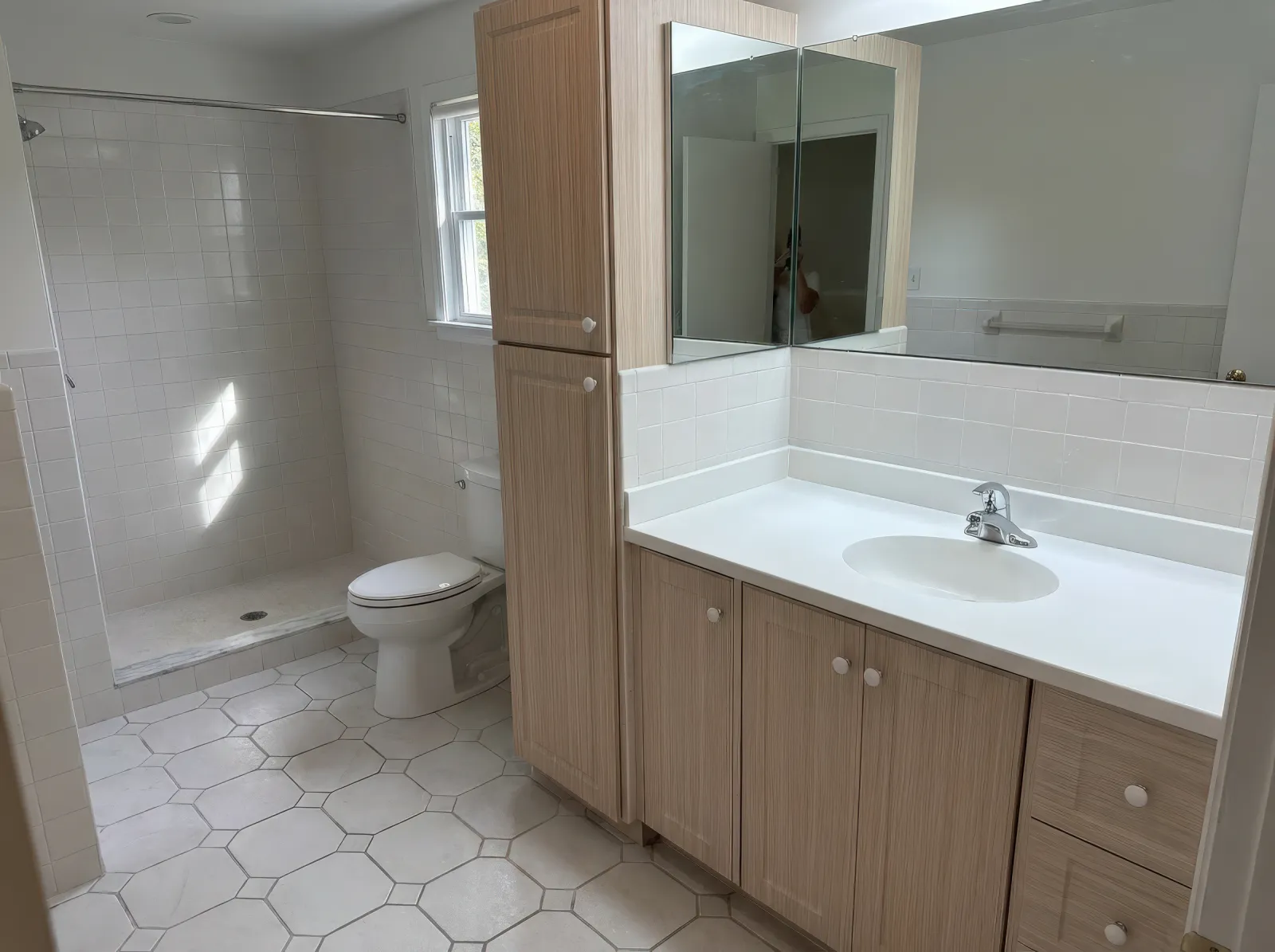 Bright bathroom with hexagonal tile floor, wooden cabinets, large mirror, shower area, and toilet near window.