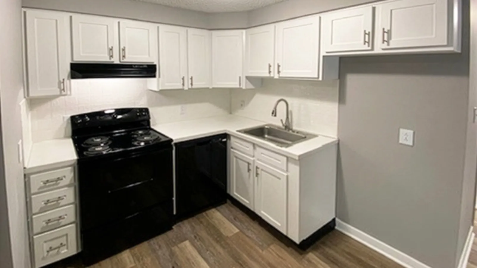 Modern kitchen with white cabinets, black appliances, stainless steel sink, and wood flooring under ceiling light.