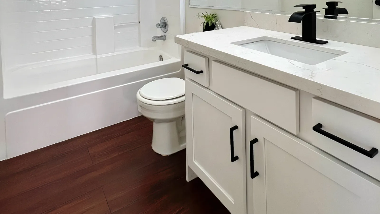 Modern bathroom with white tub, tiled wall, large mirror, black fixtures, and dark wood flooring under bright lighting.
