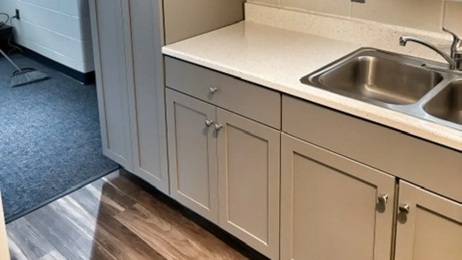 Modern gray kitchen cabinets with white countertop, stainless steel sink, and wood flooring under natural light.