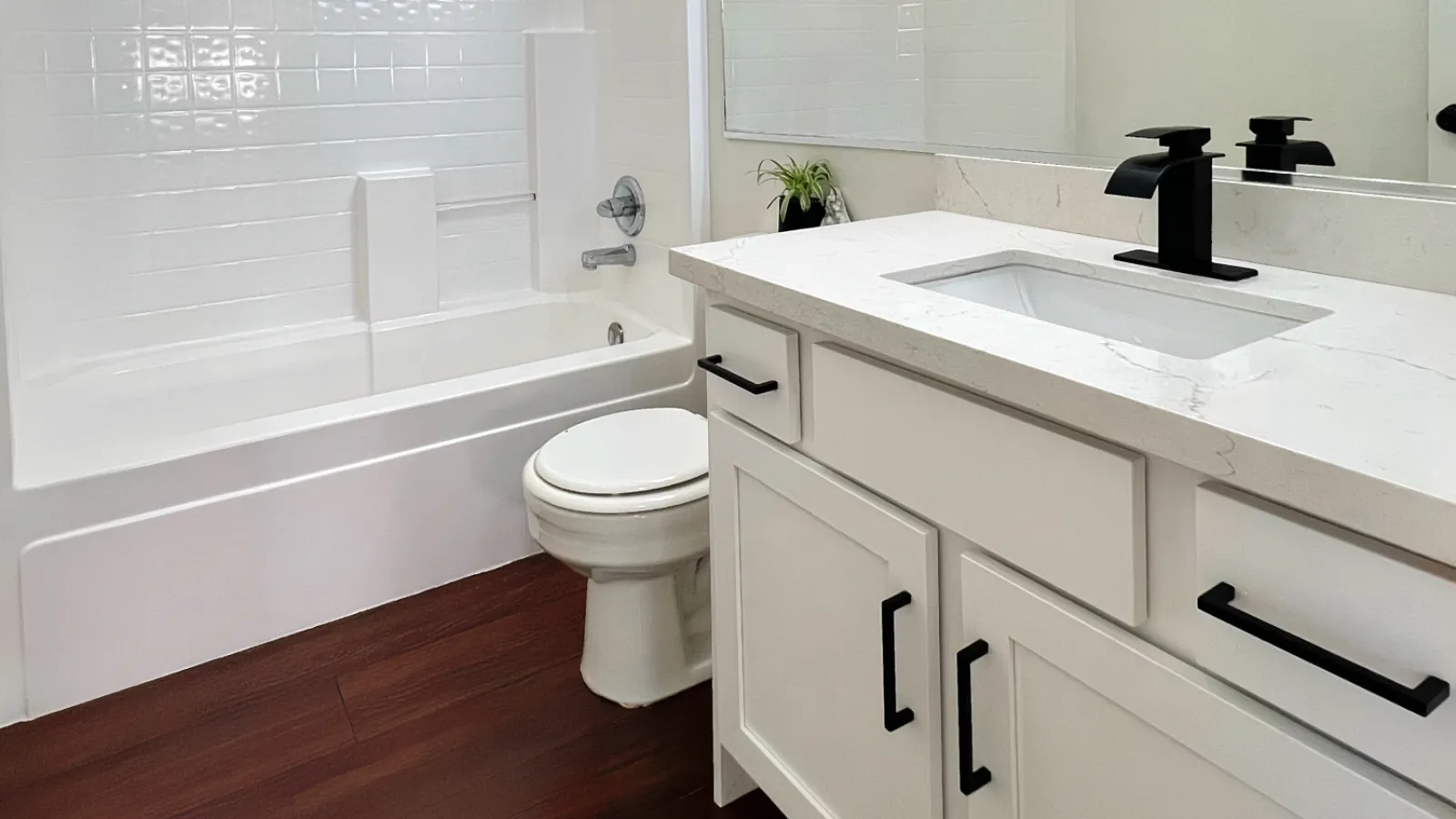 Modern bathroom with white bathtub, toilet, vanity with black fixtures, and dark wood flooring.