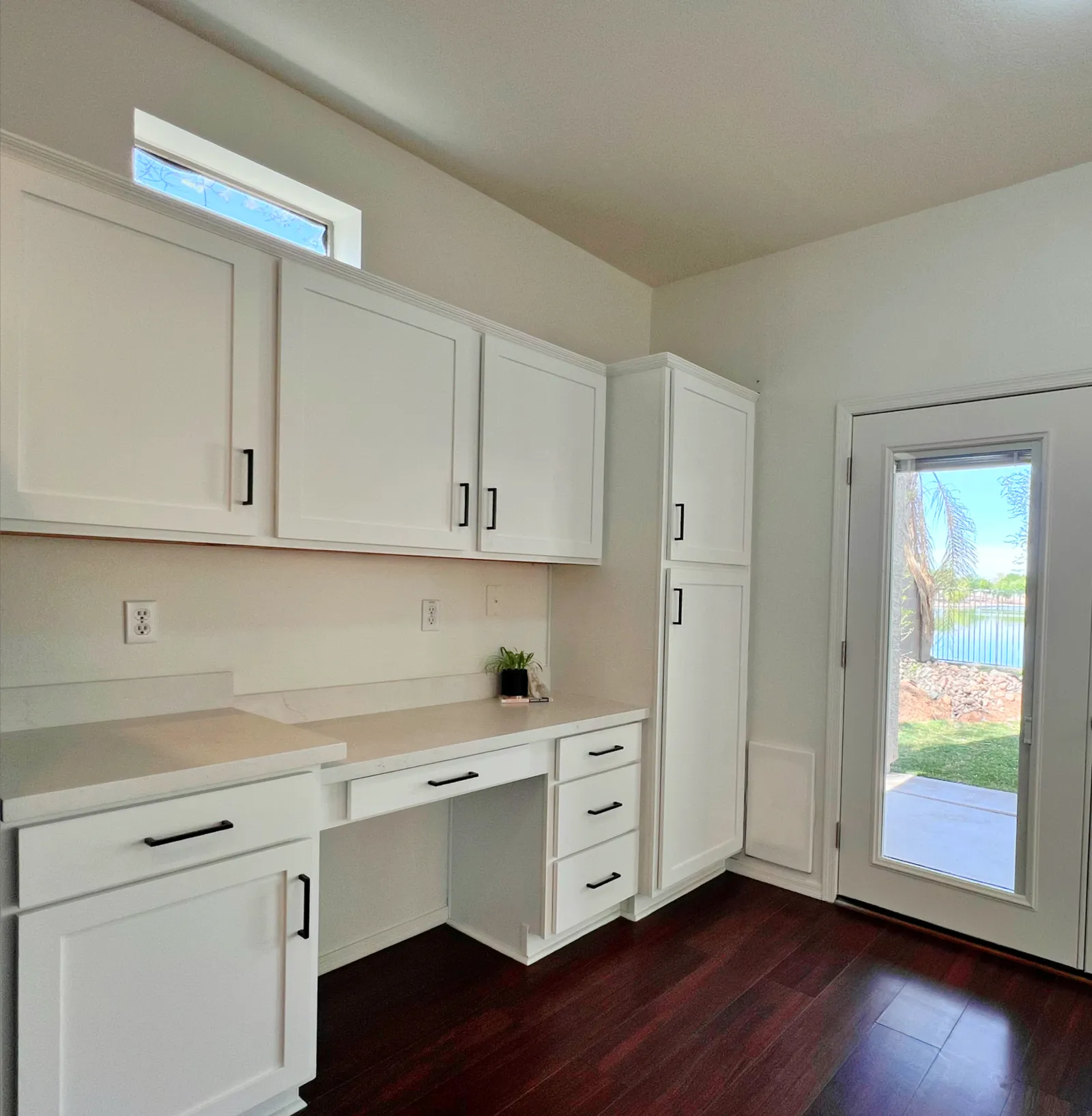 Bright white kitchen with modern cabinetry, dark wood floors, a small desk area, and a glass door overlooking a yard.