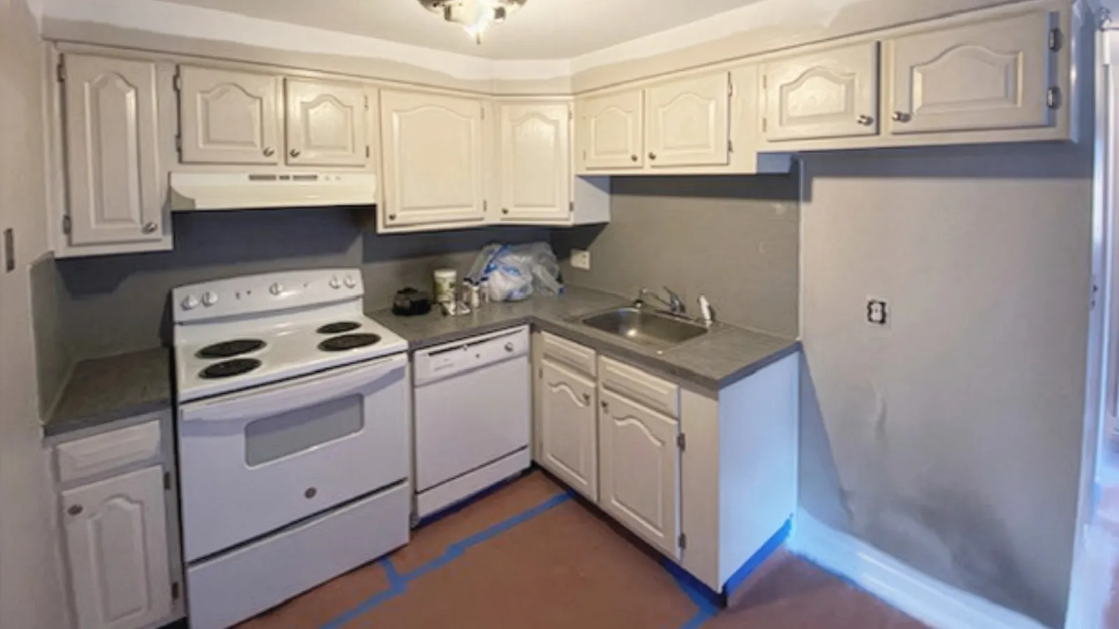 Compact kitchen with white cabinets, electric stove, dishwasher, and single sink, featuring gray countertops and walls.