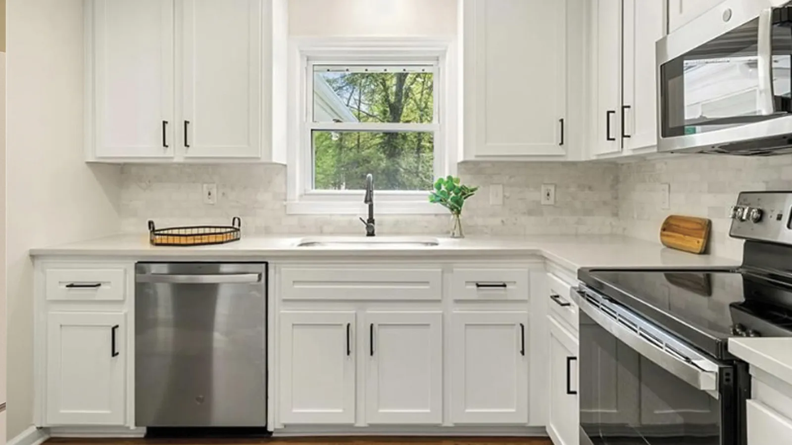 Modern white kitchen with stainless steel appliances, black hardware, and a window overlooking green trees.