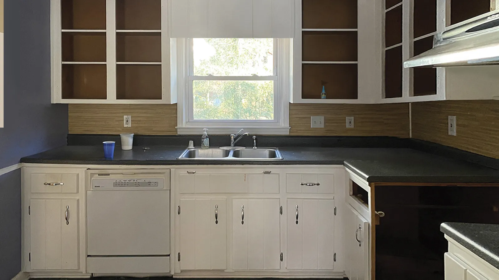 Empty kitchen with white cabinets, black countertops, dishwasher, and window above double sink under daylight.