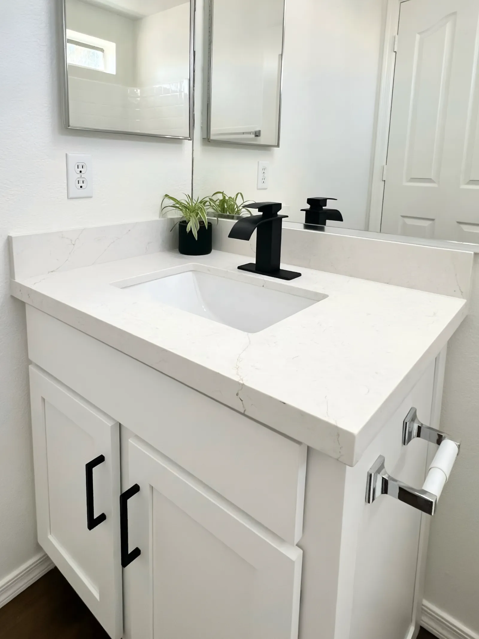 Modern white bathroom vanity with marble countertop, black faucet, small green plant, and rectangular mirror.