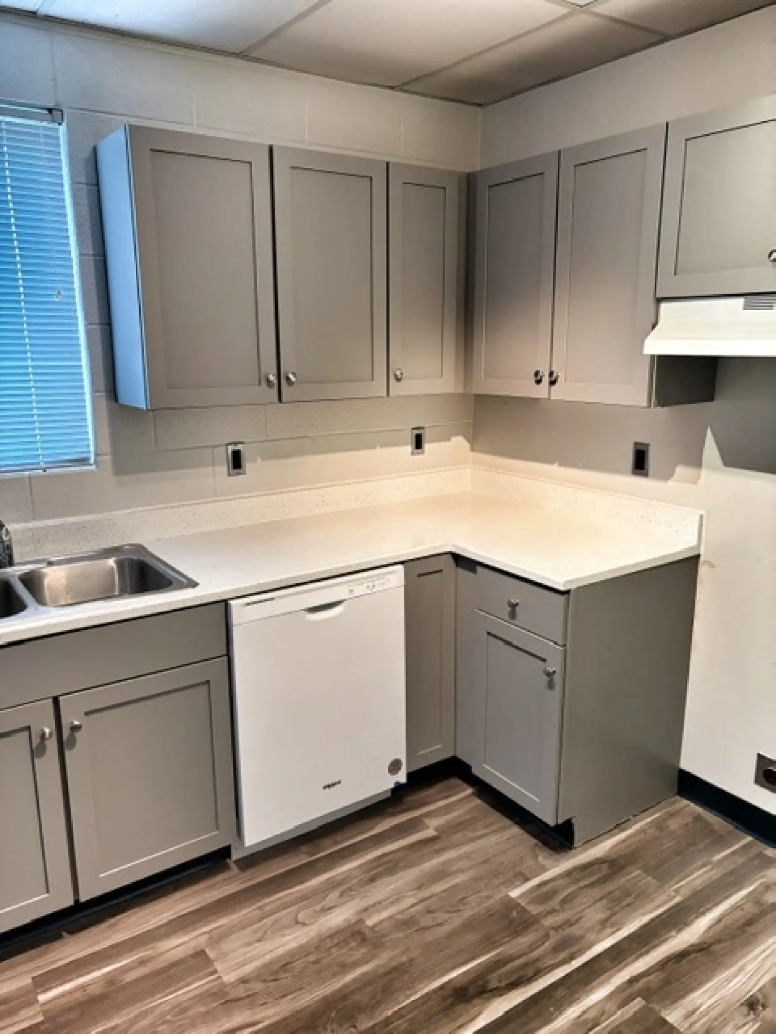 Modern kitchen corner with gray cabinets, white countertop, dishwasher, stainless steel sink, and wood laminate floor.