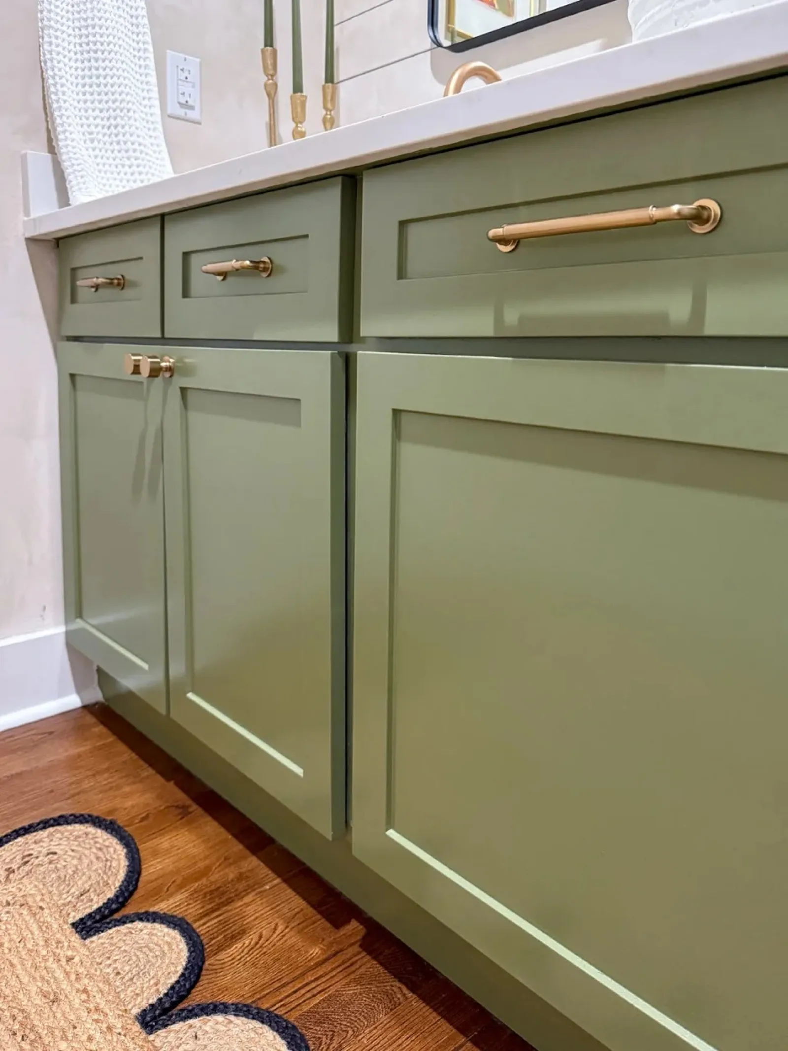 Close-up of olive green kitchen cabinets with brass handles and hardwood flooring with woven rug edge.