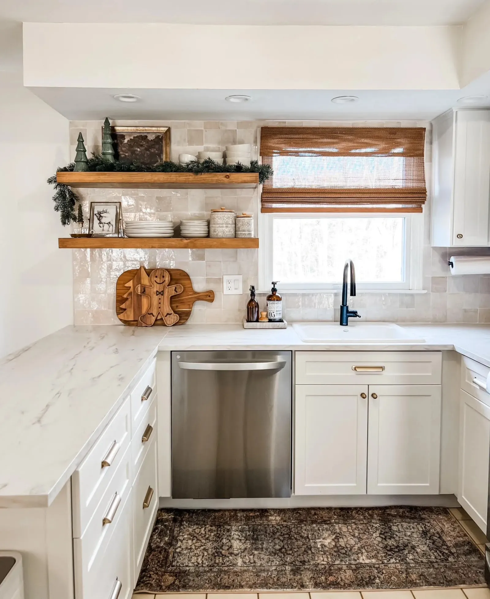 Bright kitchen with white cabinets, stainless steel dishwasher, wooden shelves, festive decor, and a window with a bamboo shade.
