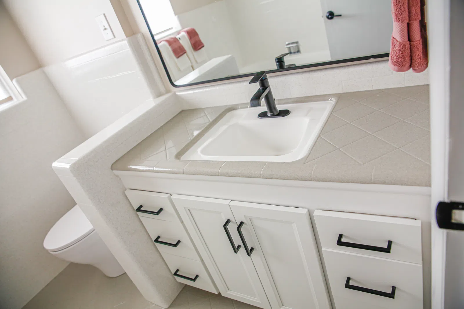 Modern bathroom vanity with white cabinets, black fixtures, and a beige tiled countertop next to a white toilet.