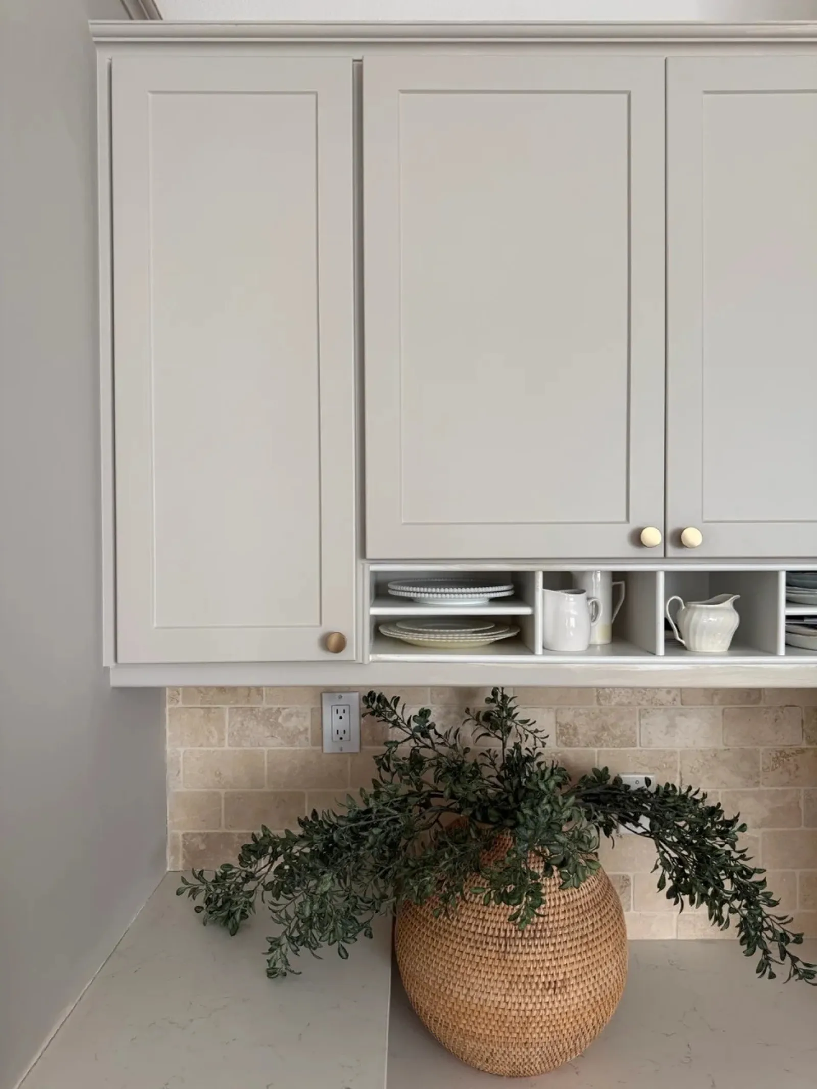 Modern kitchen with beige cabinets, open shelves holding dishes, and a wicker vase with greenery on marble countertop.