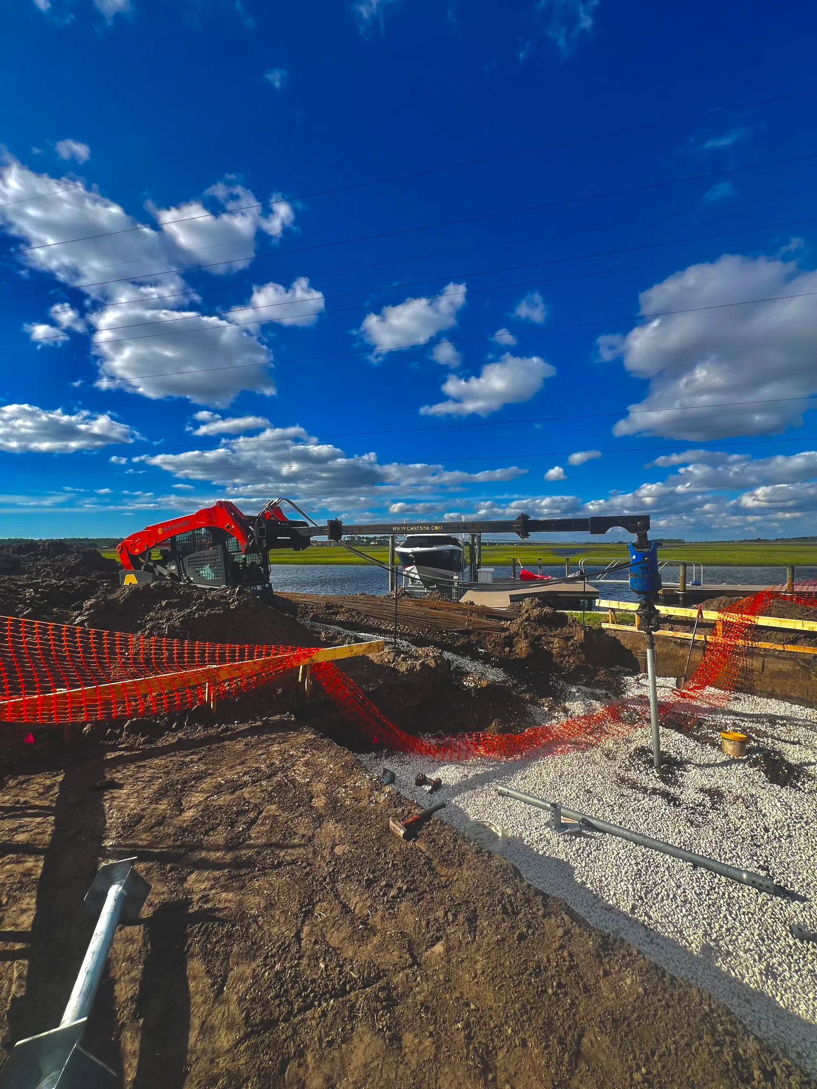 Construction site by water with a red excavator, orange safety netting, gravel, and blue sky with clouds.