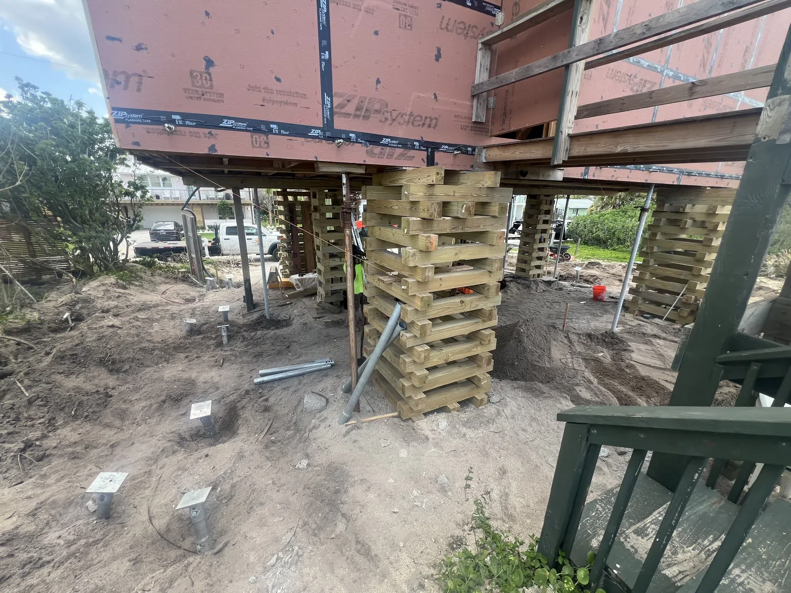 House elevated on layered wooden supports with metal piers in sandy soil during construction.