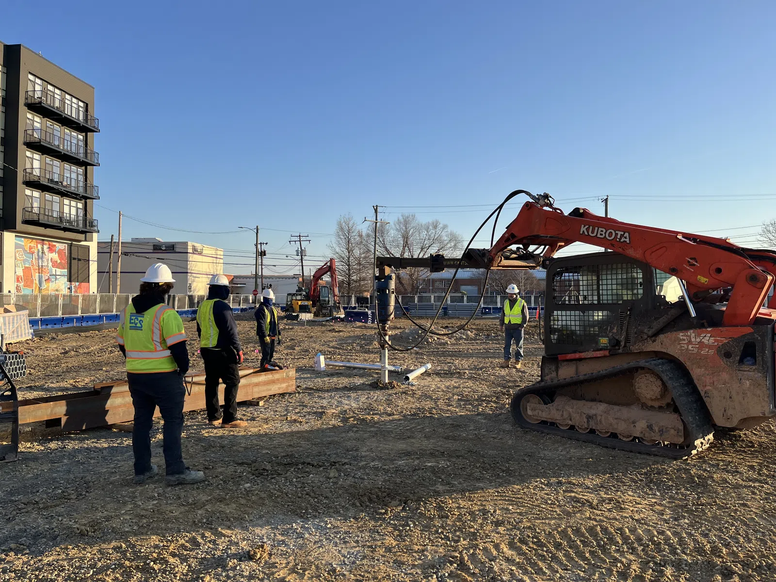 Construction workers in safety gear operating Kubota machinery on a sunny building site with urban backdrop.