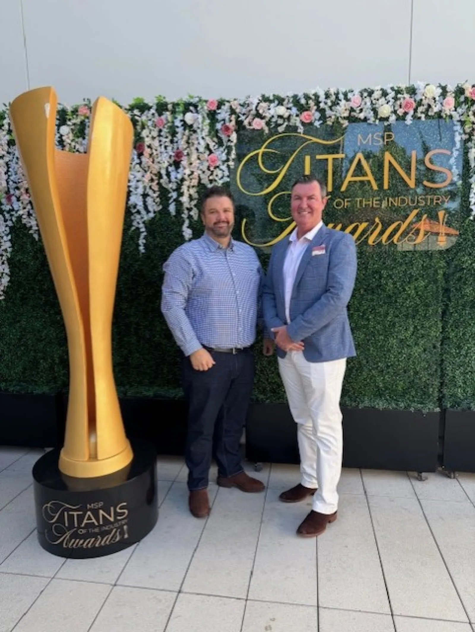 Two men posing beside a large golden award trophy at the MSP Titans of the Industry Awards event with floral backdrop.