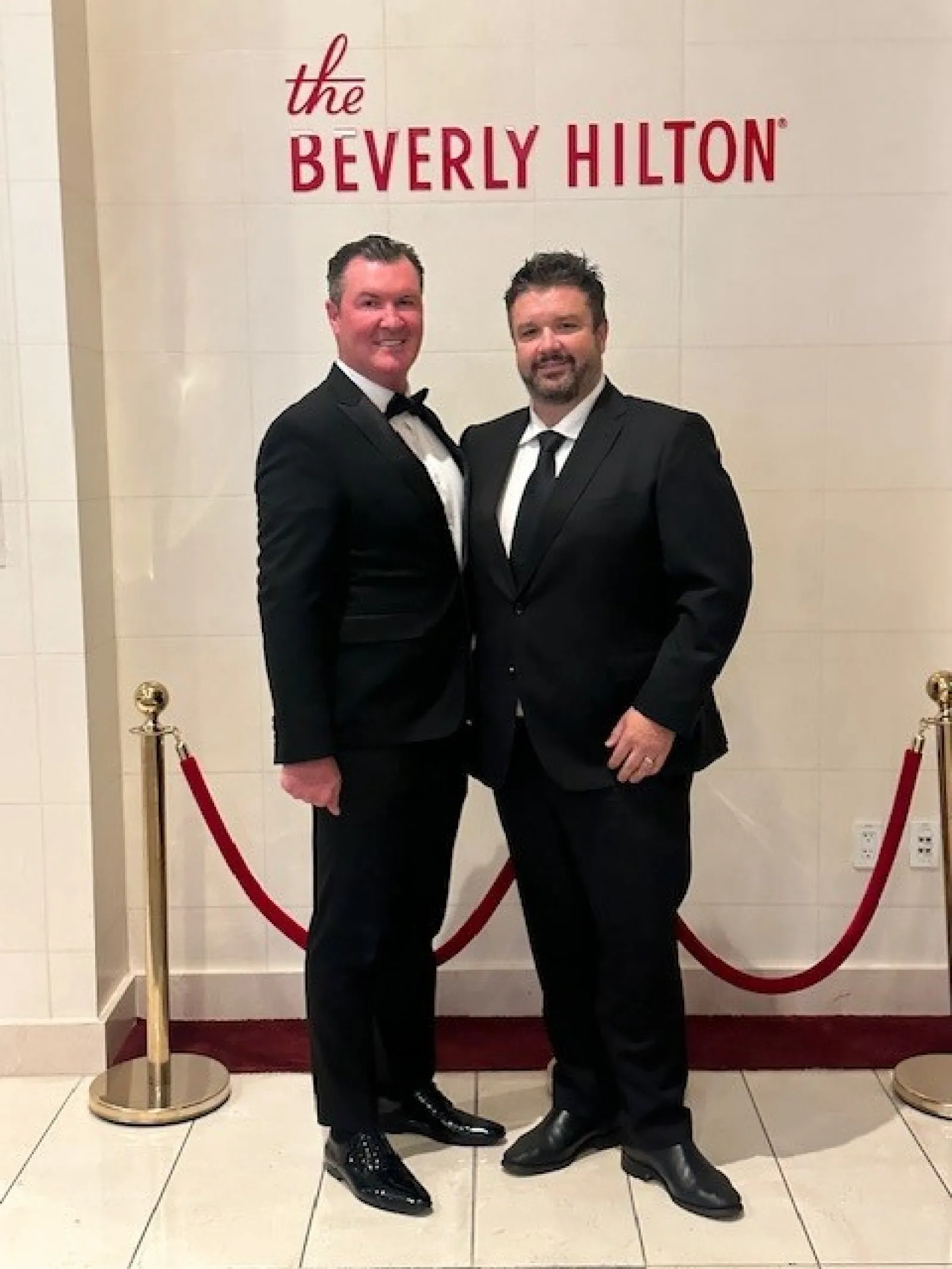 Two men in black tuxedos posing in front of a Beverly Hilton wall with red velvet ropes around them.