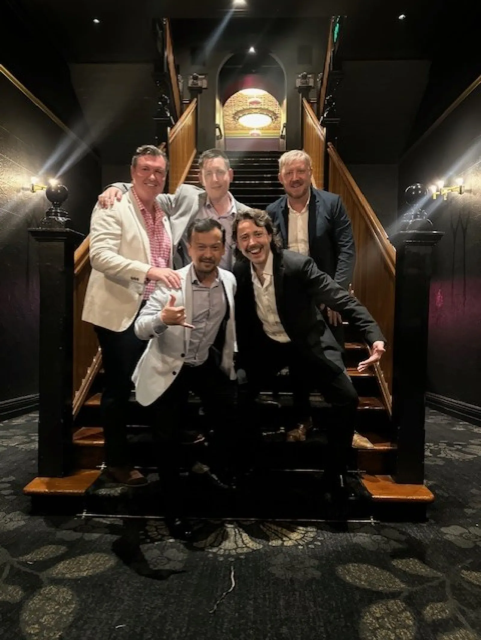 Five men in formal attire posing on wooden staircase inside a dimly lit elegant building with carpeted floor.