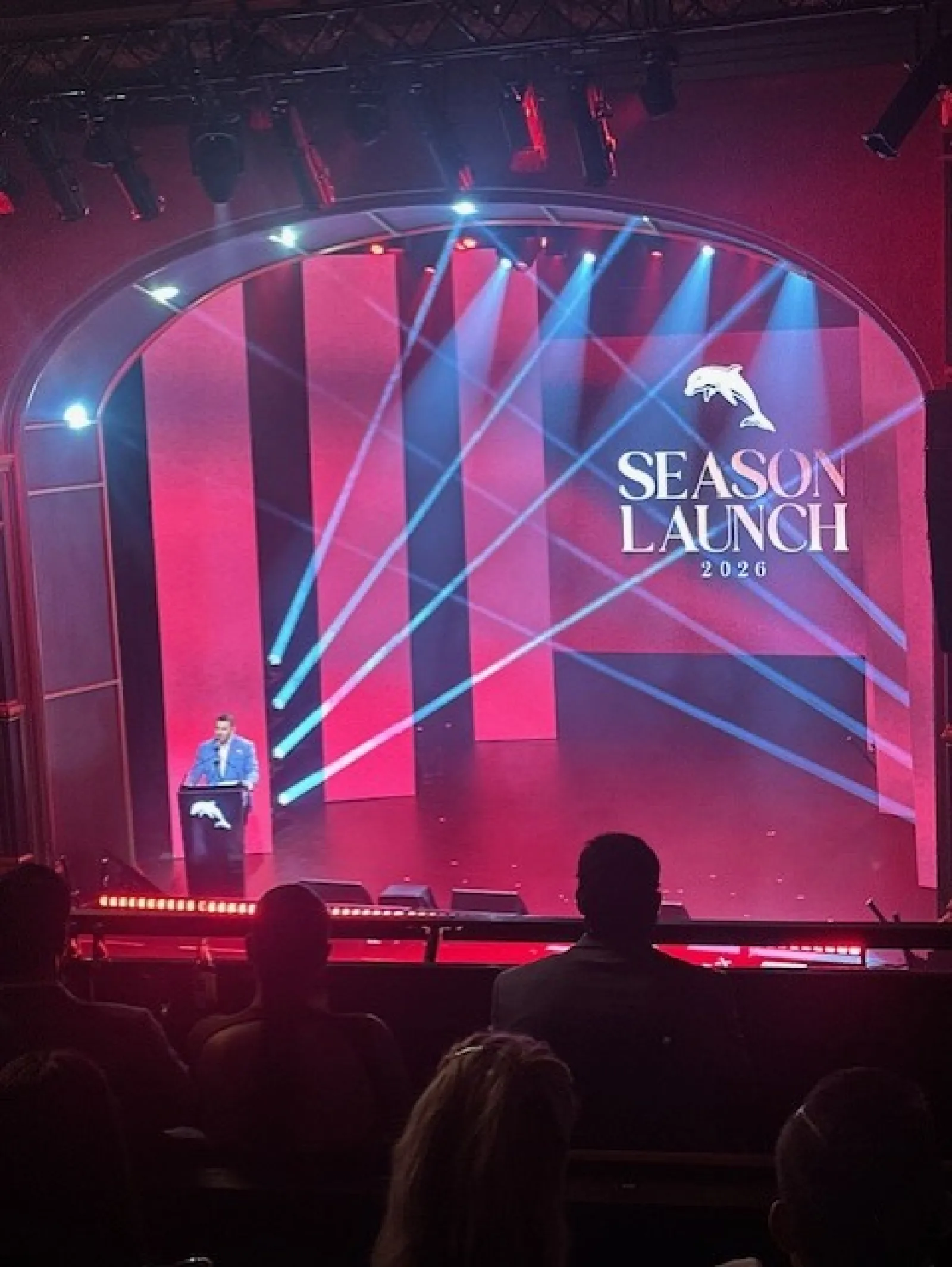 Speaker at podium on stage with red panels and blue spotlight beams during 2026 Season Launch event