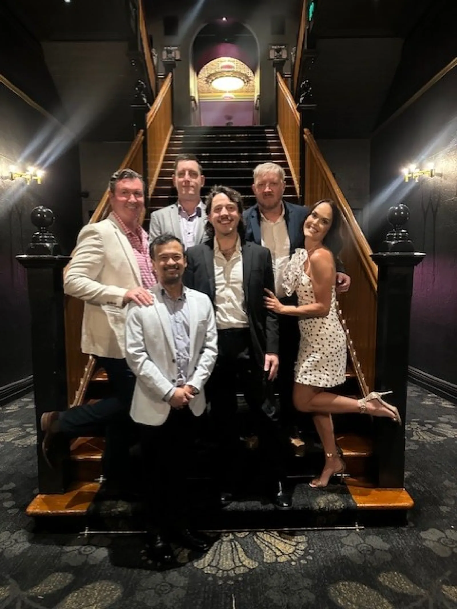 Group of six people dressed formally posing by a grand staircase in an elegant indoor setting.