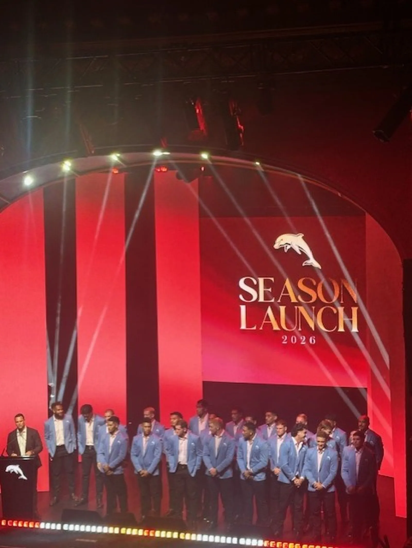 Group of men in blue blazers standing on stage at Season Launch 2026 event with red background and dolphin logo.