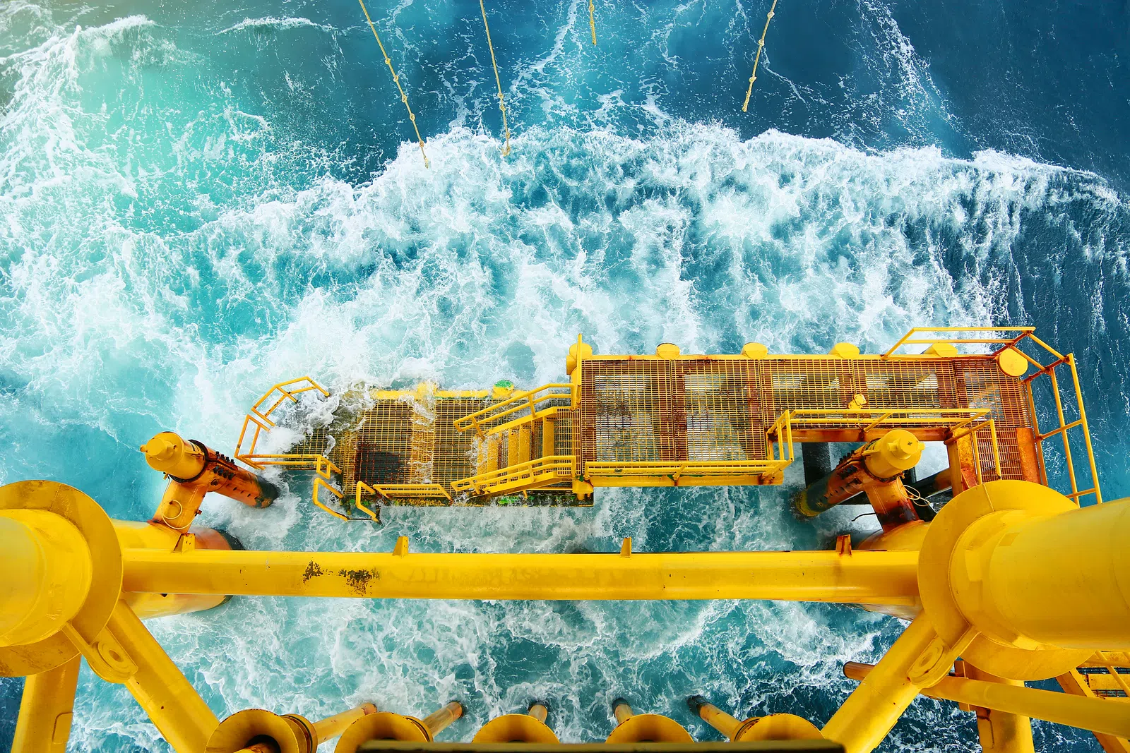 Top-down view of yellow offshore oil rig platform over blue ocean waves with foamy white surf below.