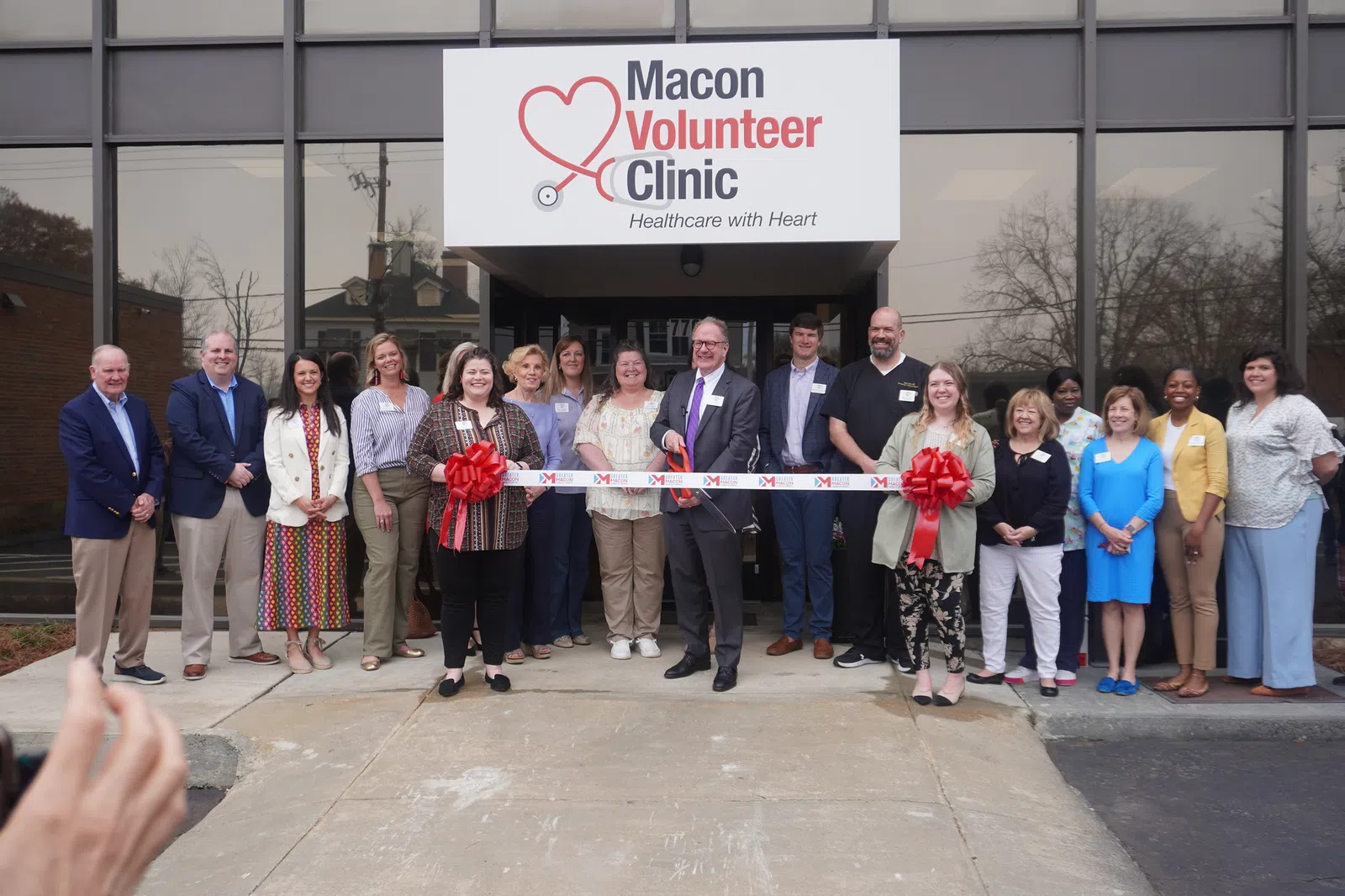 Group of people at ribbon-cutting event outside Macon Volunteer Clinic with overlaid text on sign above entrance.