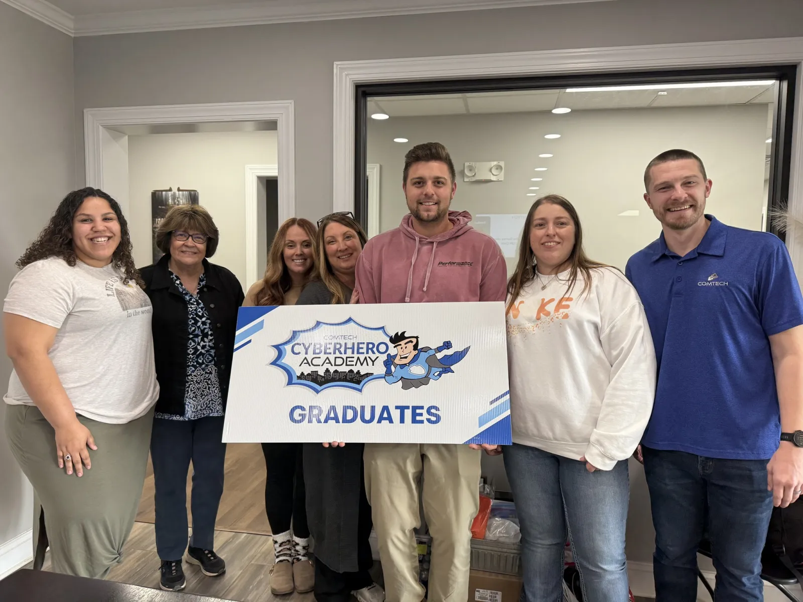 Group of seven people smiling and holding a sign that reads Cyberhero Academy Graduates inside an office.