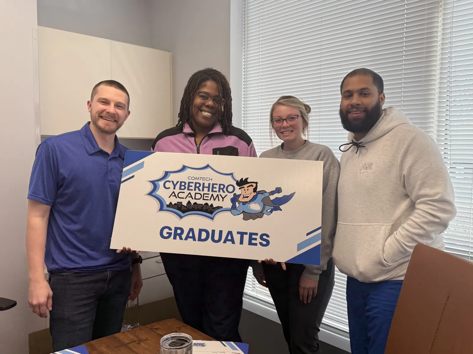 Four diverse graduates smiling and holding a ComTec CyberHero Academy Graduates sign in an office setting.