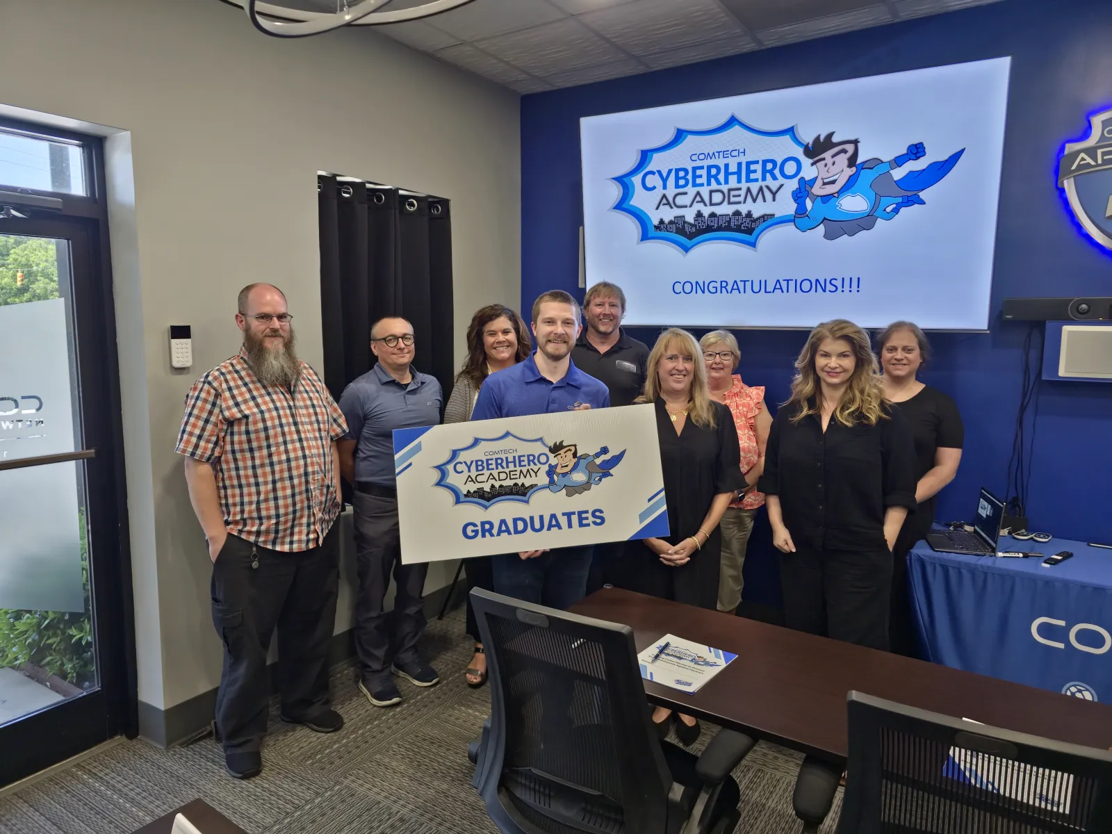 Group of diverse graduates holding CyberHero Academy sign in office celebrating completion.