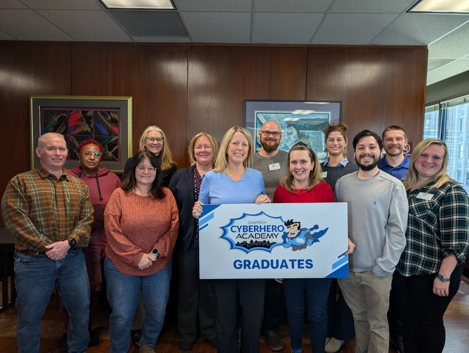 Group of diverse graduates holding a Cyberhero Academy sign in an office setting with wooden walls and framed art.