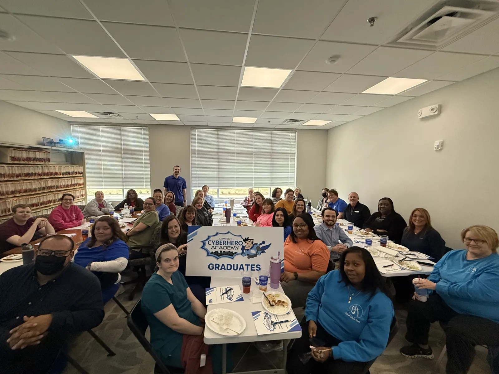Group of Cyberhero Academy graduates seated around tables in a bright room holding a graduates sign.