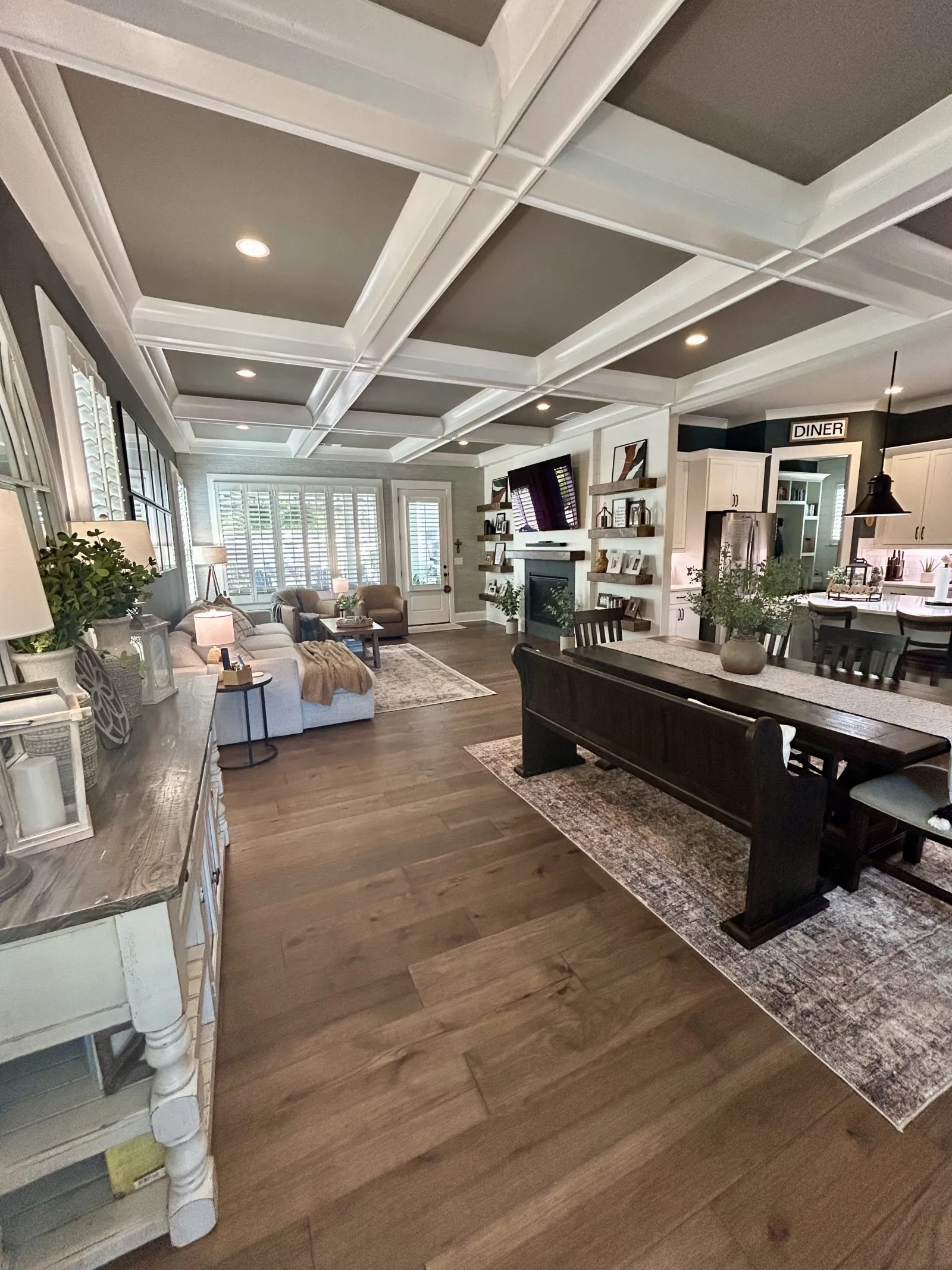 Open-concept living and dining room with hardwood floors, coffered ceiling, white cabinetry, and farmhouse decor.