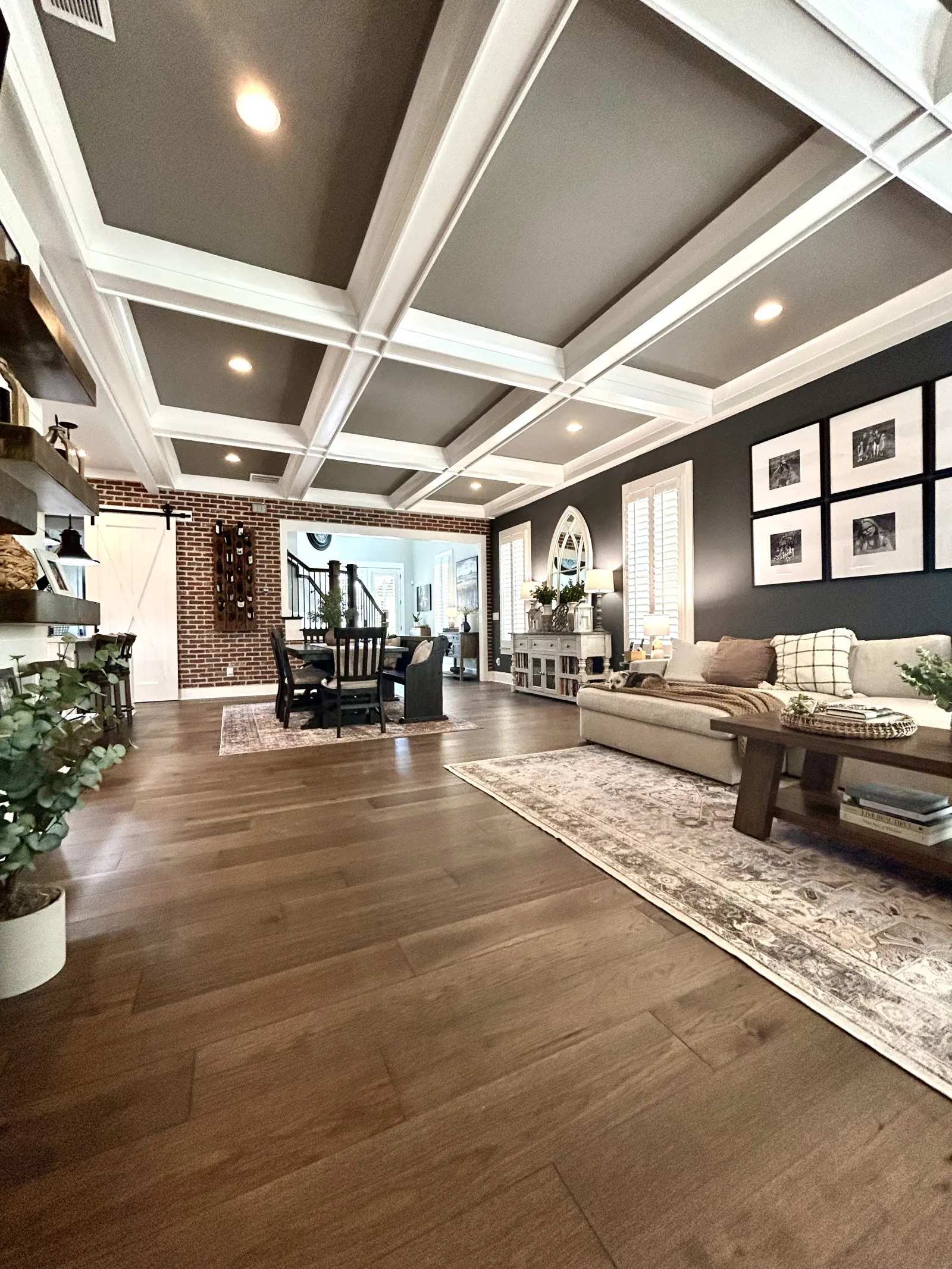 Spacious living room with coffered ceiling, hardwood floors, sectional sofa, dining area, and dark accent wall with framed photos.