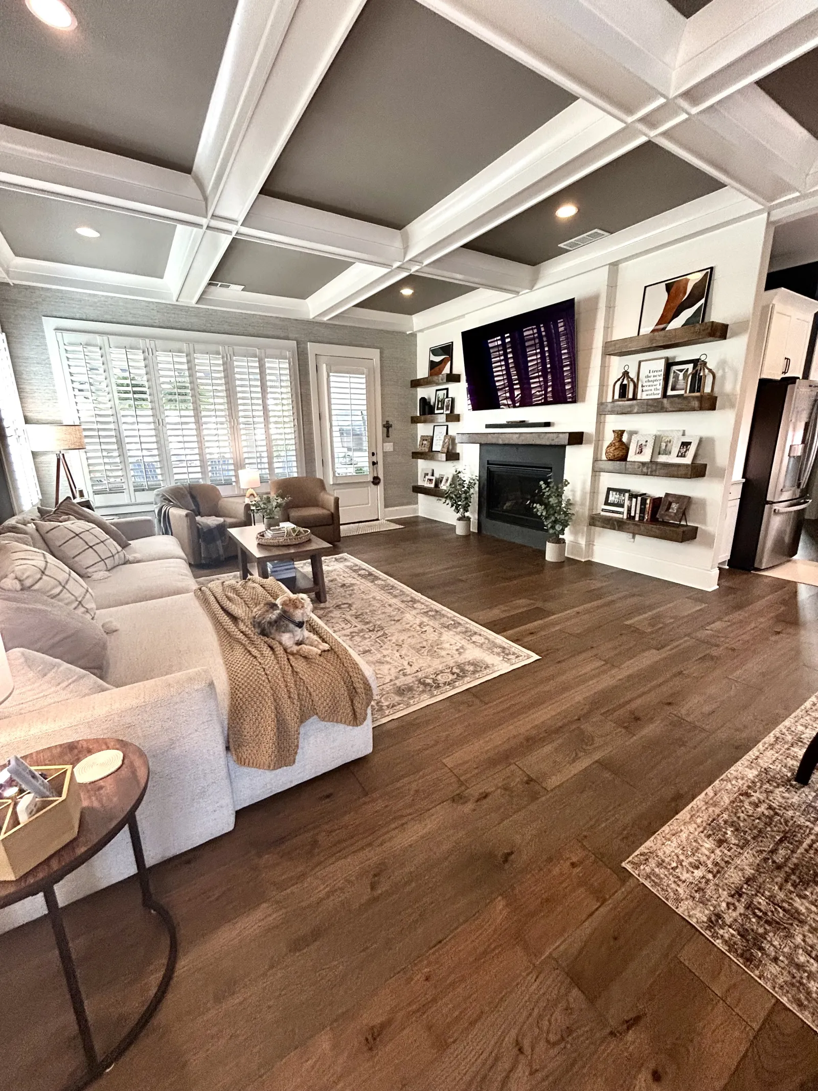 Cozy living room with beige sofa, armchairs, wooden flooring, fireplace, mounted TV, and decorative shelves.