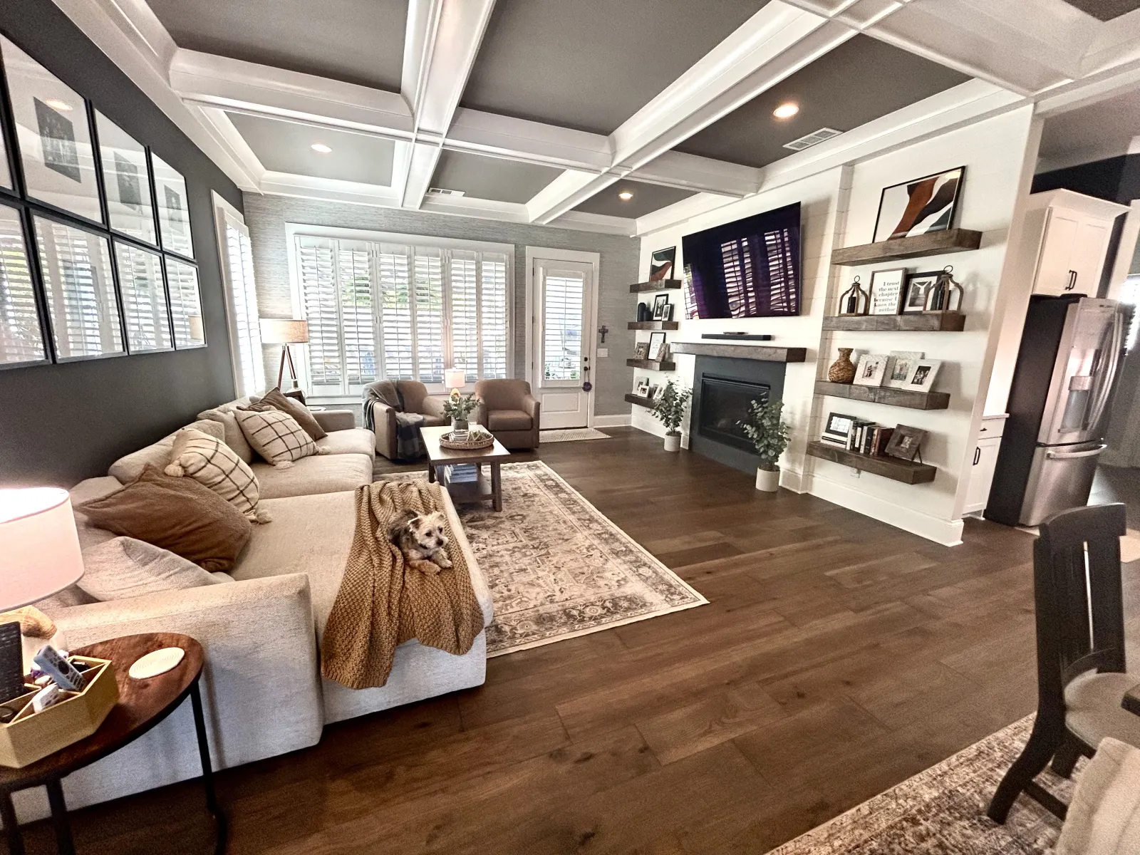 Cozy modern living room with beige sofa, wooden floors, fireplace, TV, and large windows with white shutters.