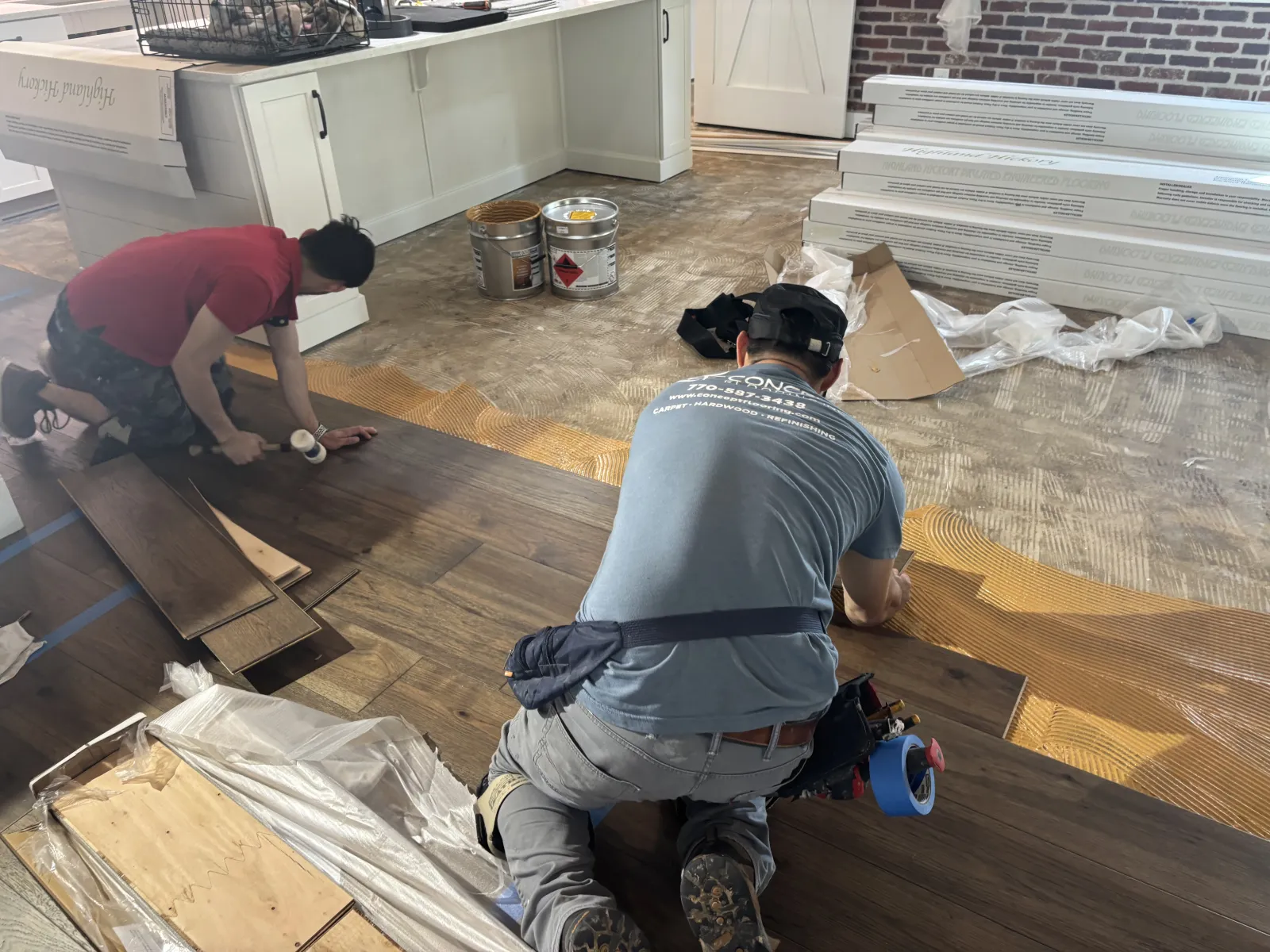 Two workers installing dark wood flooring on a kitchen floor with tools and supplies around them.