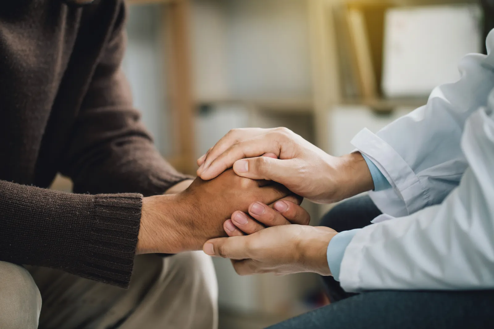 Close up image of a medical professional in a white coat holding the hands of a patient in a brown sweater.